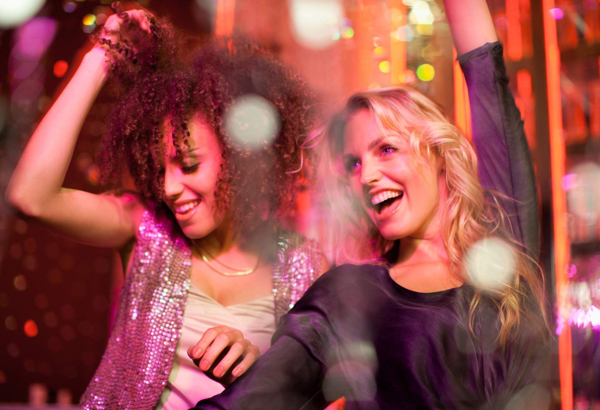A blonde and a curly-haired woman smile while dancing and raising their hands in the air.