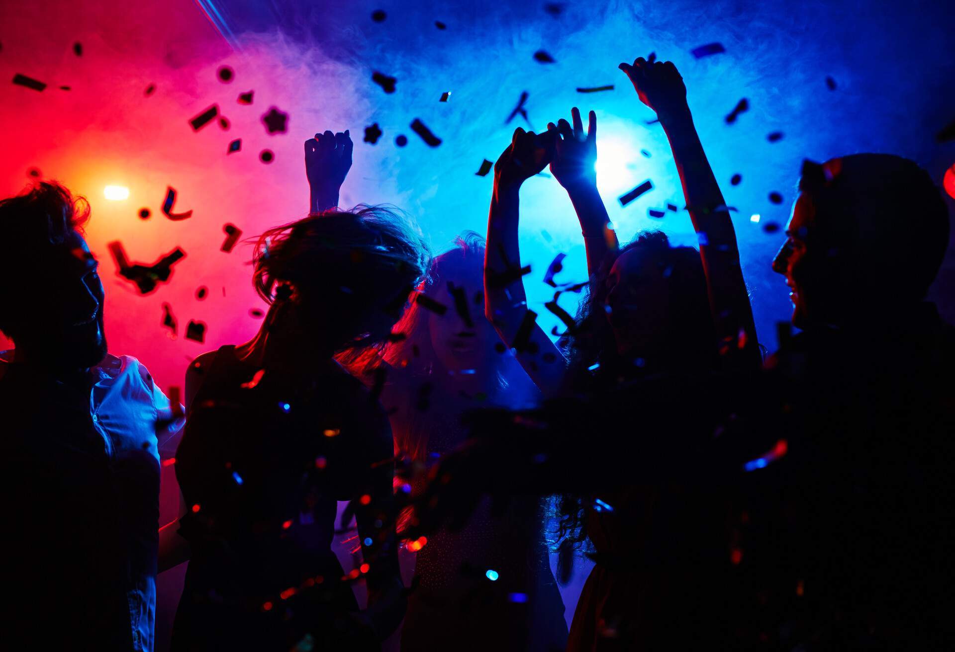 Silhouette of people dancing with their hands in the air against the colourful lights and confetti.