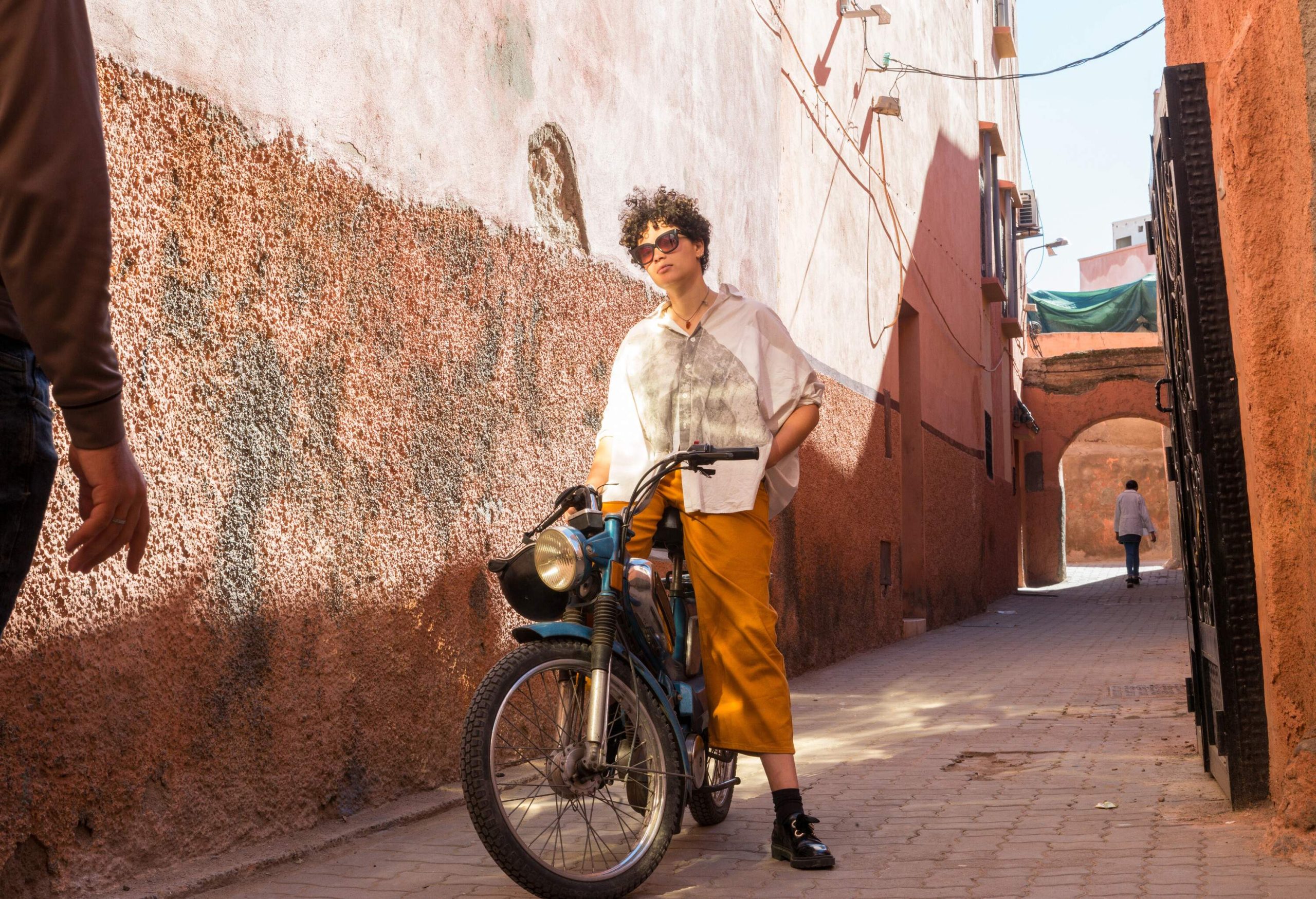A short, curly-haired lady poses in a blue motorcycle on a cobbled alleyway.