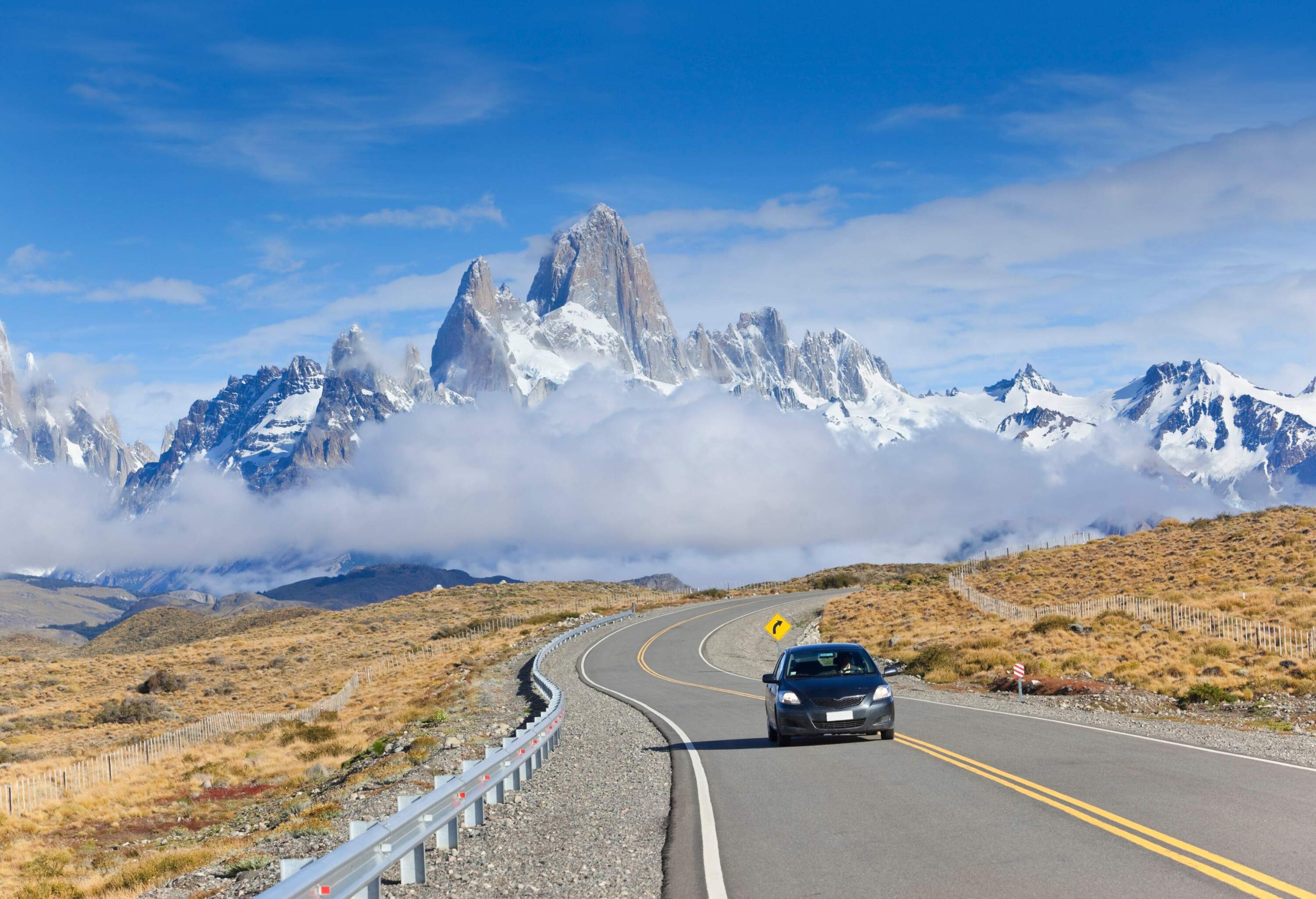 Rising triumphantly in the background, the iconic Mount Fitz Roy, a striking granite peak that towers over the Patagonian landscape, shrouded in mist, creates a dramatic and unforgettable scene as a car navigates the winding road away from this natural wonder.