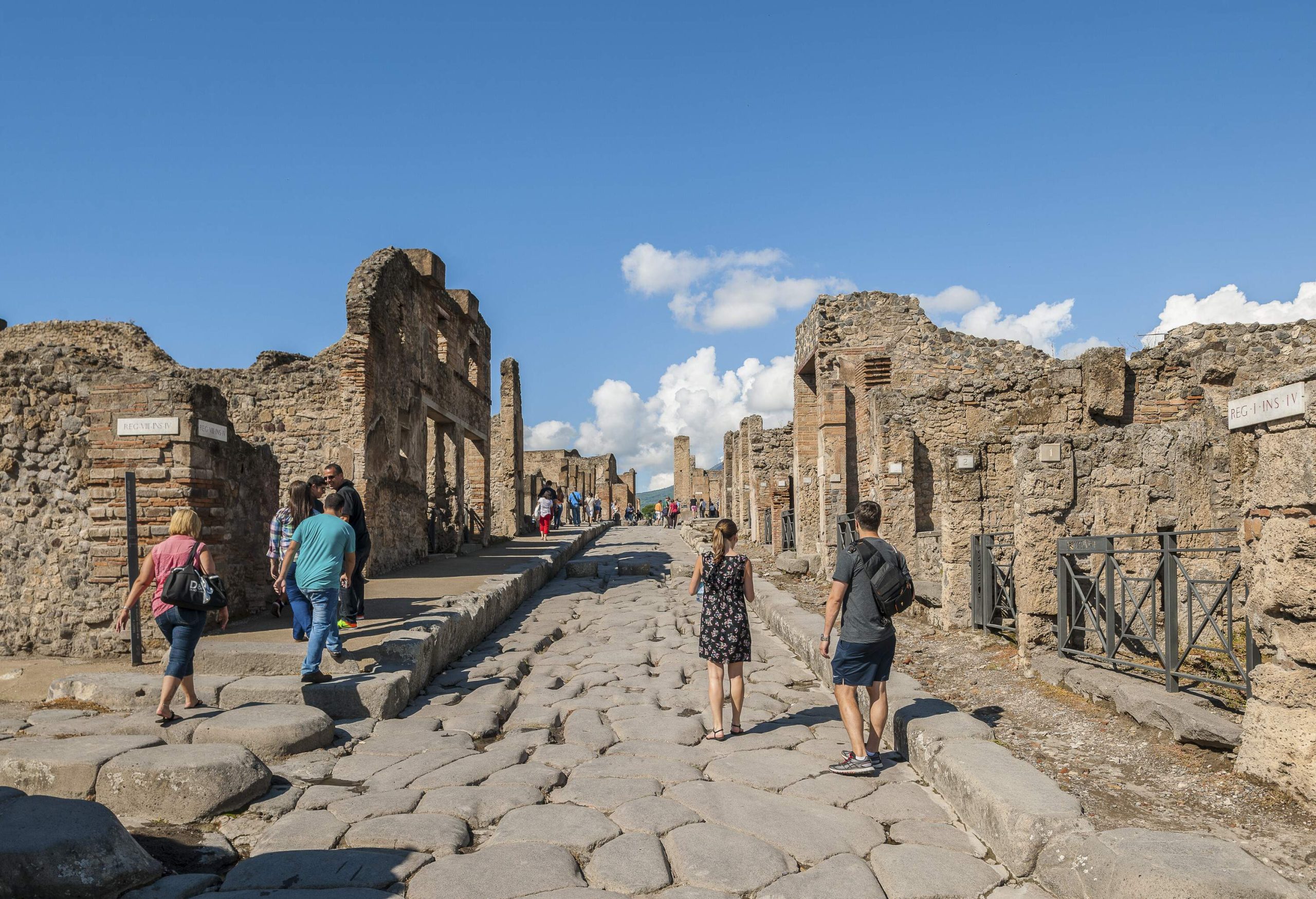 Tourists stroll on a cobblestone street along the ruins of an archaeological site.