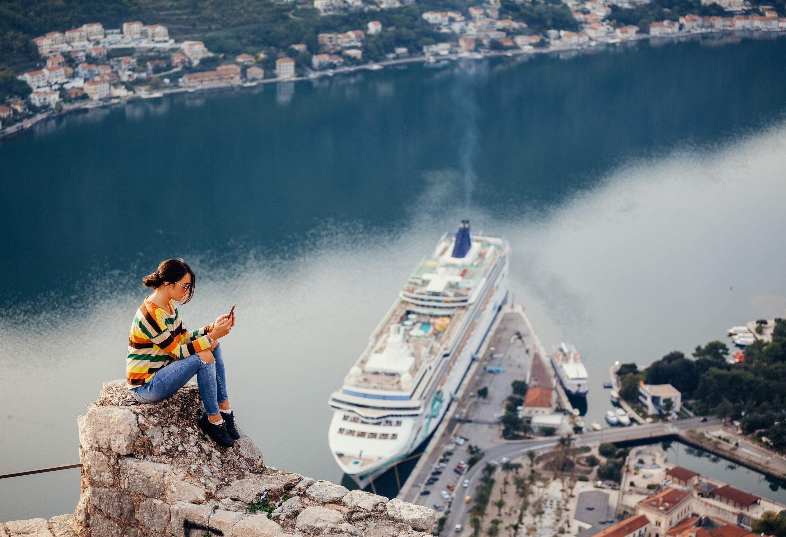 A young woman using a smartphone while sitting on a ledge overlooking a big ship docked in a harbour.