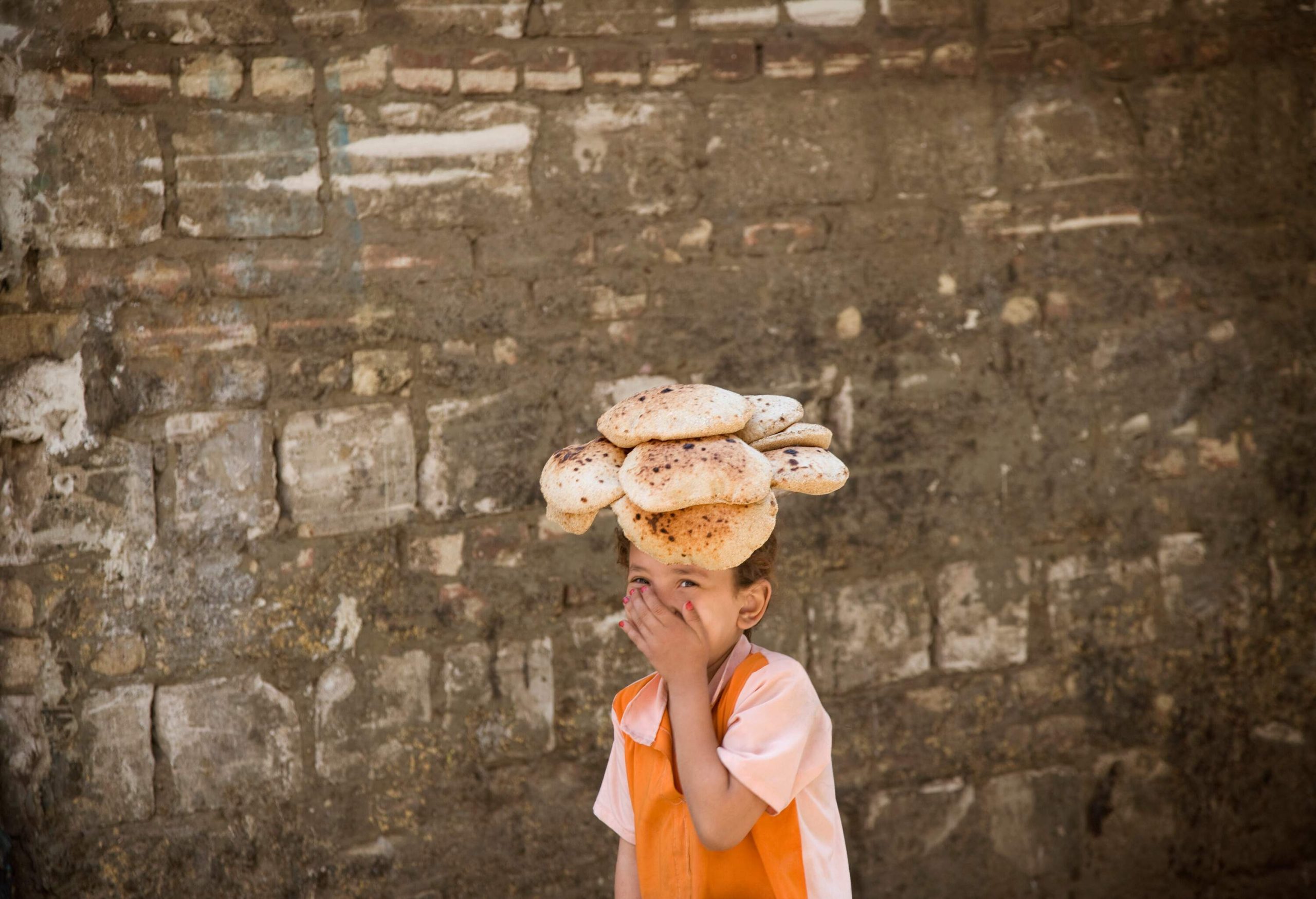 A girl smiles and covers her mouth as she carries a bundle of bread on her head.