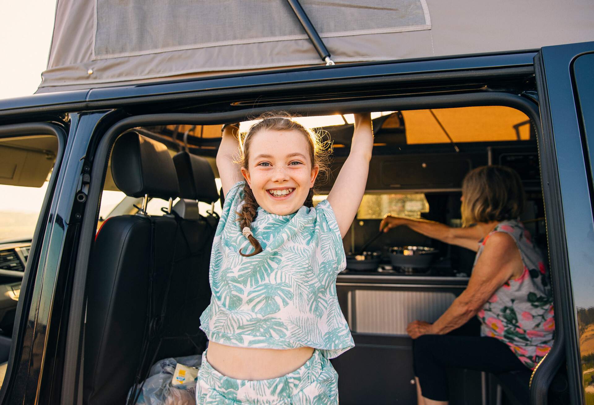 A happy young girl smiling from her camper van. In the background, her grandmother is preparing dinner. 