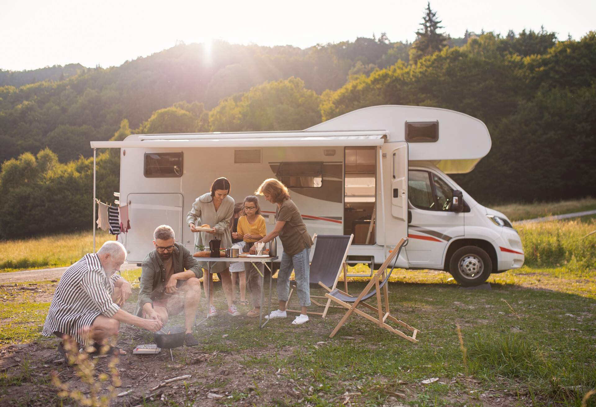 A multi-generation family sitting and eating outdoors by car, caravan holiday trip. 