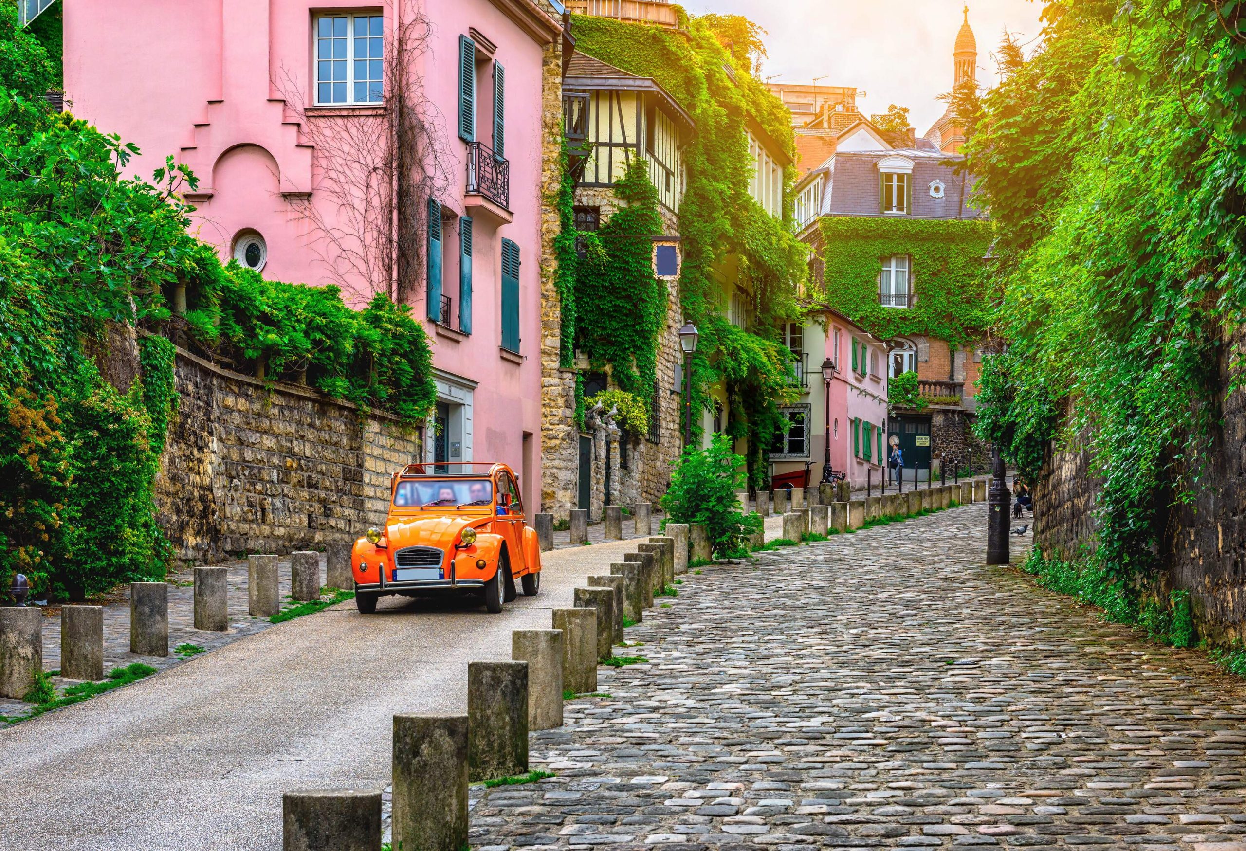 An orange vintage car running down a steep road along a cobbled sidewalk and colourful houses.