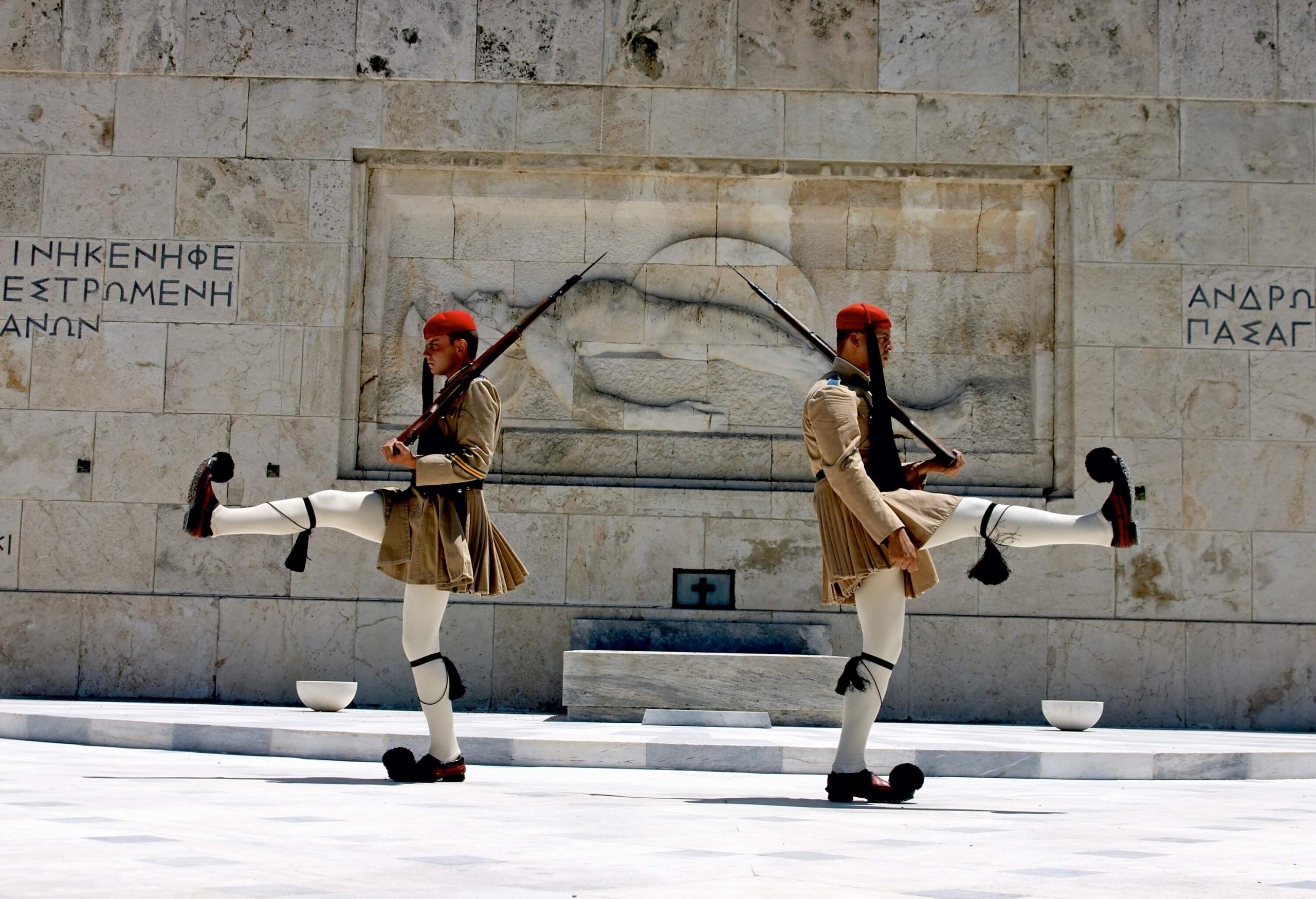 Two elite Greek soldiers wearing traditional Evzones uniforms back to back, with firearms on their left shoulders and their right legs up.