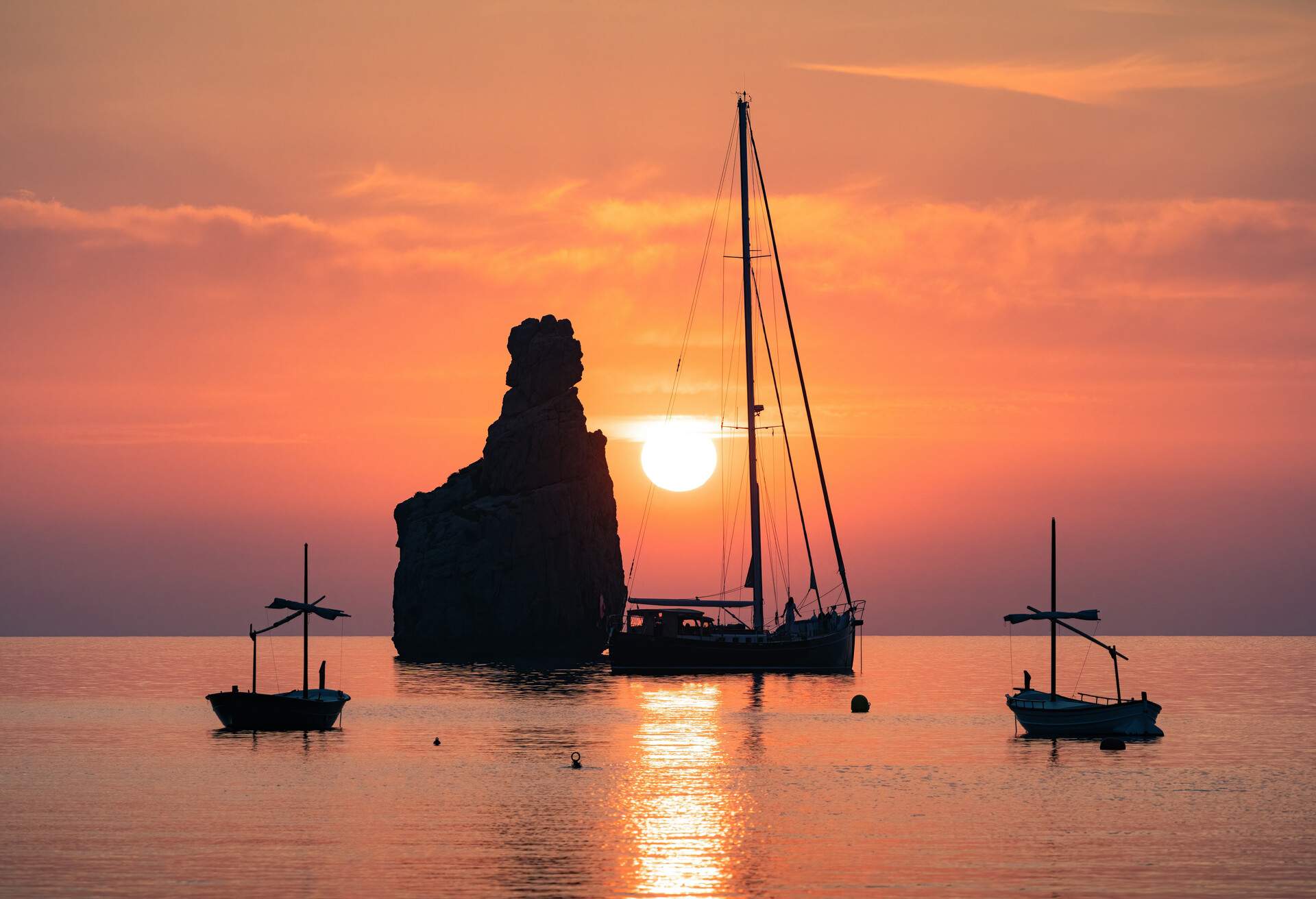 A big rock that lies in the middle of the bay, surrounded by anchored boats over the calm water at sunset.