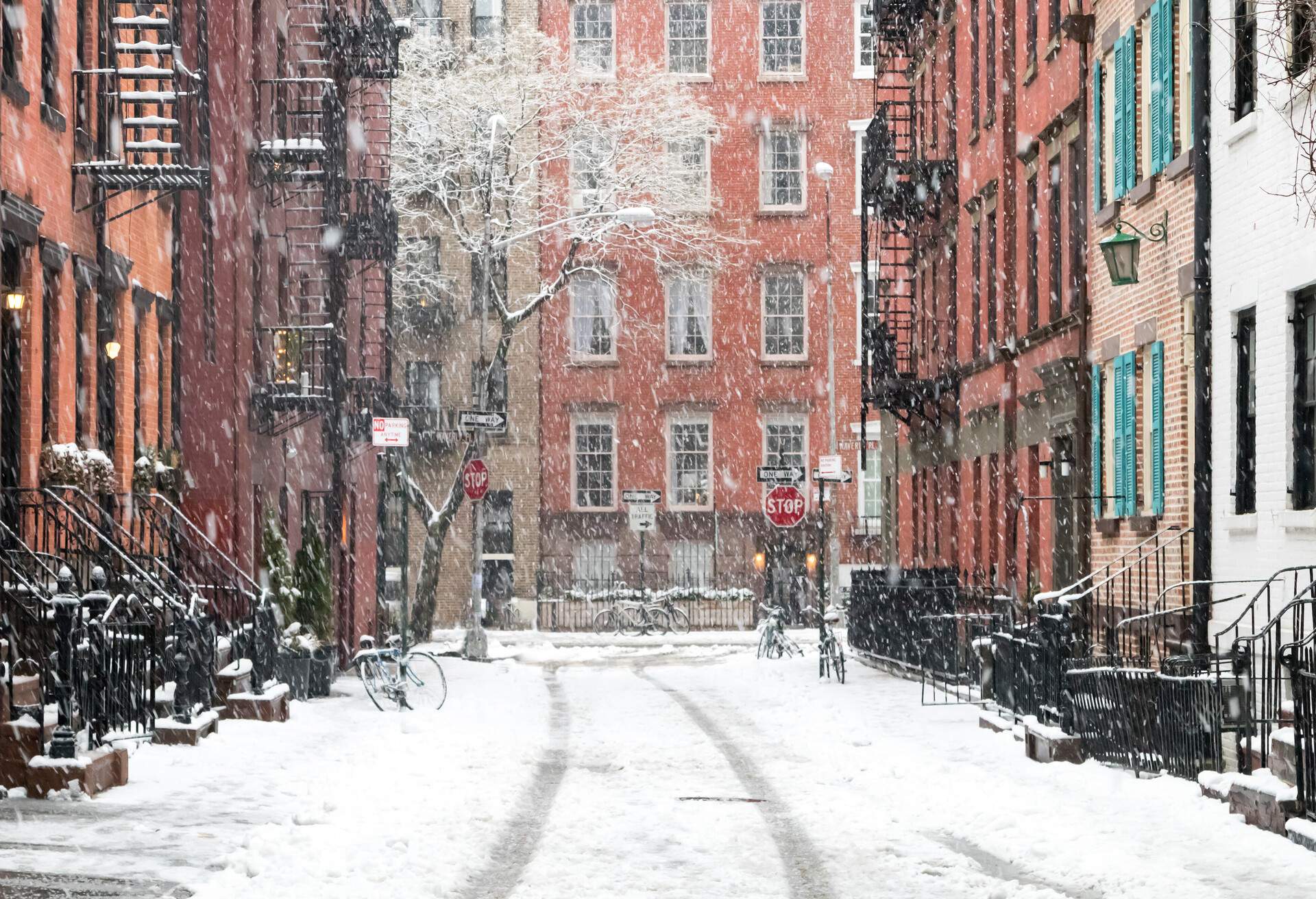 Tire prints on a snow path bordered by adjacent colourful brick buildings during a snowfall.