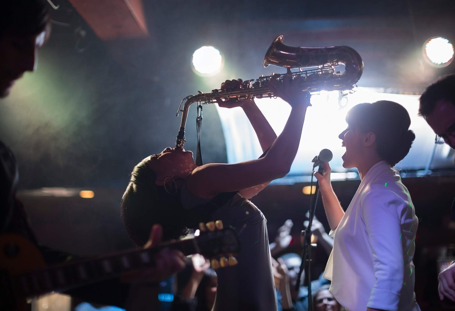 A person sings into a microphone while a musician plays a saxophone while bending backwards.