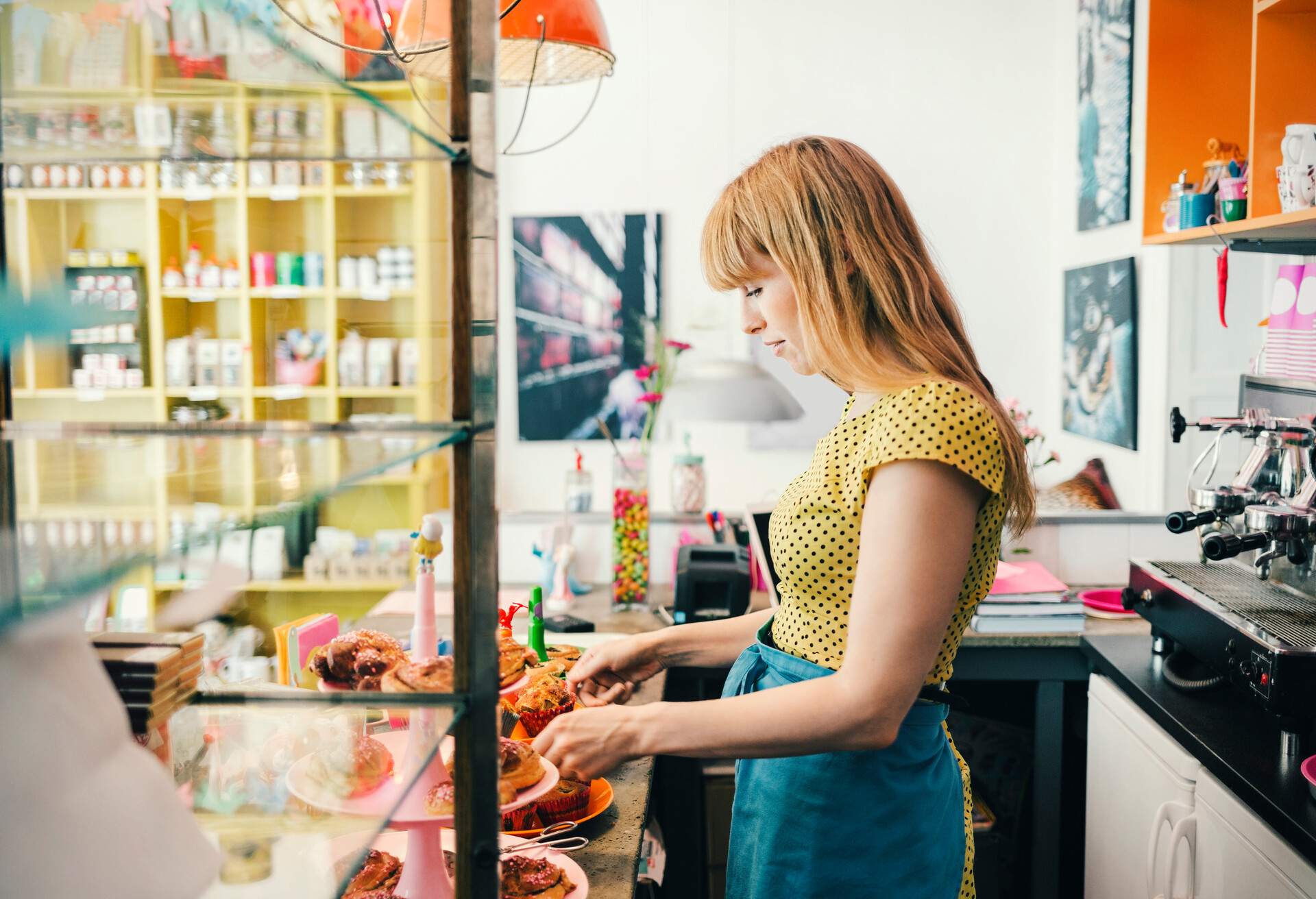 A woman in a yellow shirt prepares an assortment of pastries on a store counter.