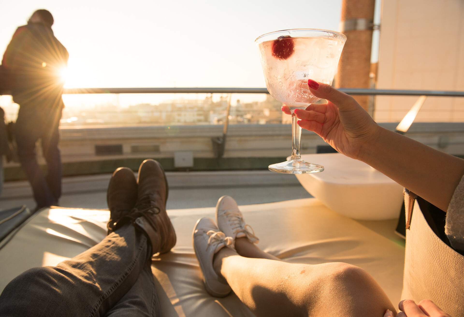 Girl raises cocktail glass to toast as she lies on the sofa of a rooftop bar during sunset in Madrid, Spain