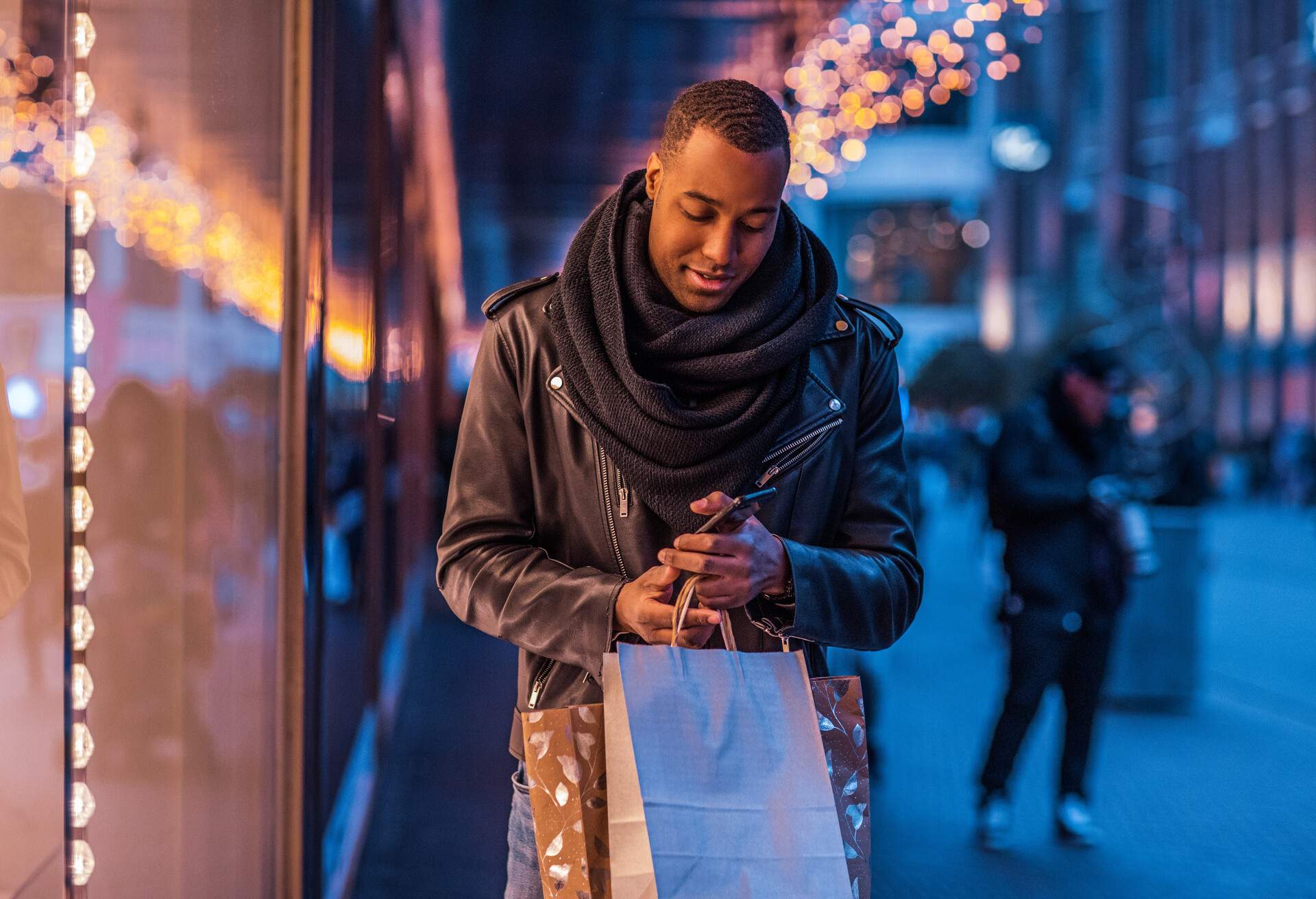 A male holding his smartphone and paper bags walks on the sidewalk along the shops.