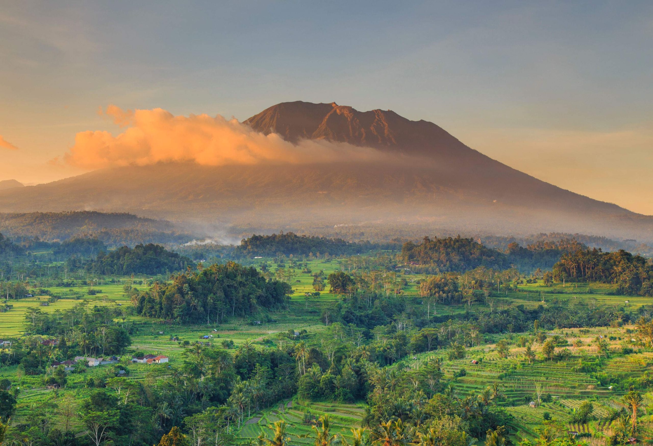 An awe-inspiring volcano with a cloud-kissed peak stands in a gorgeous natural landscape with terrace fields cascading down the hills and lush trees dotting the landscape.