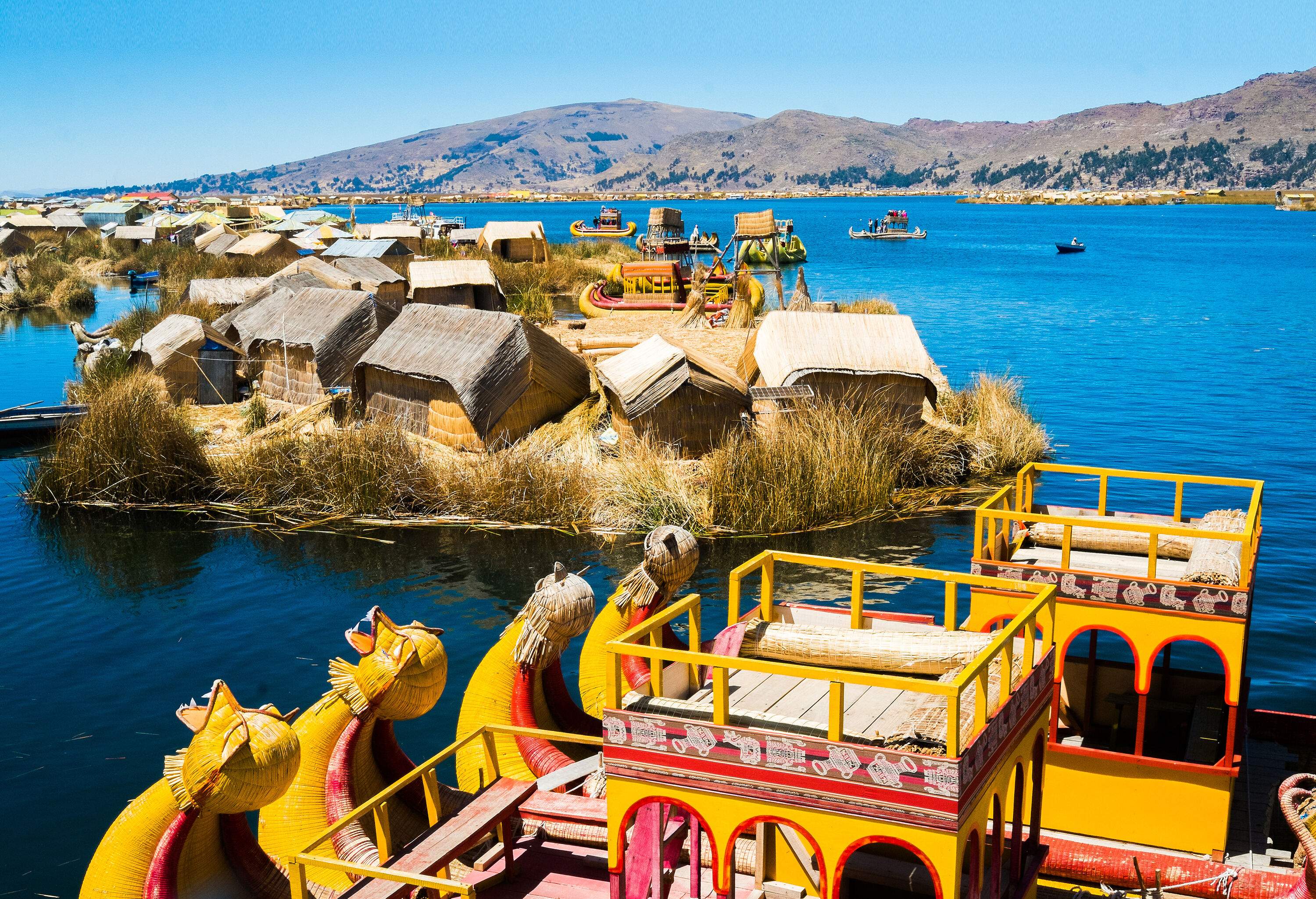 Houses on floating islands with traditional boats surrounded by a lake with rolling hills in the distance.