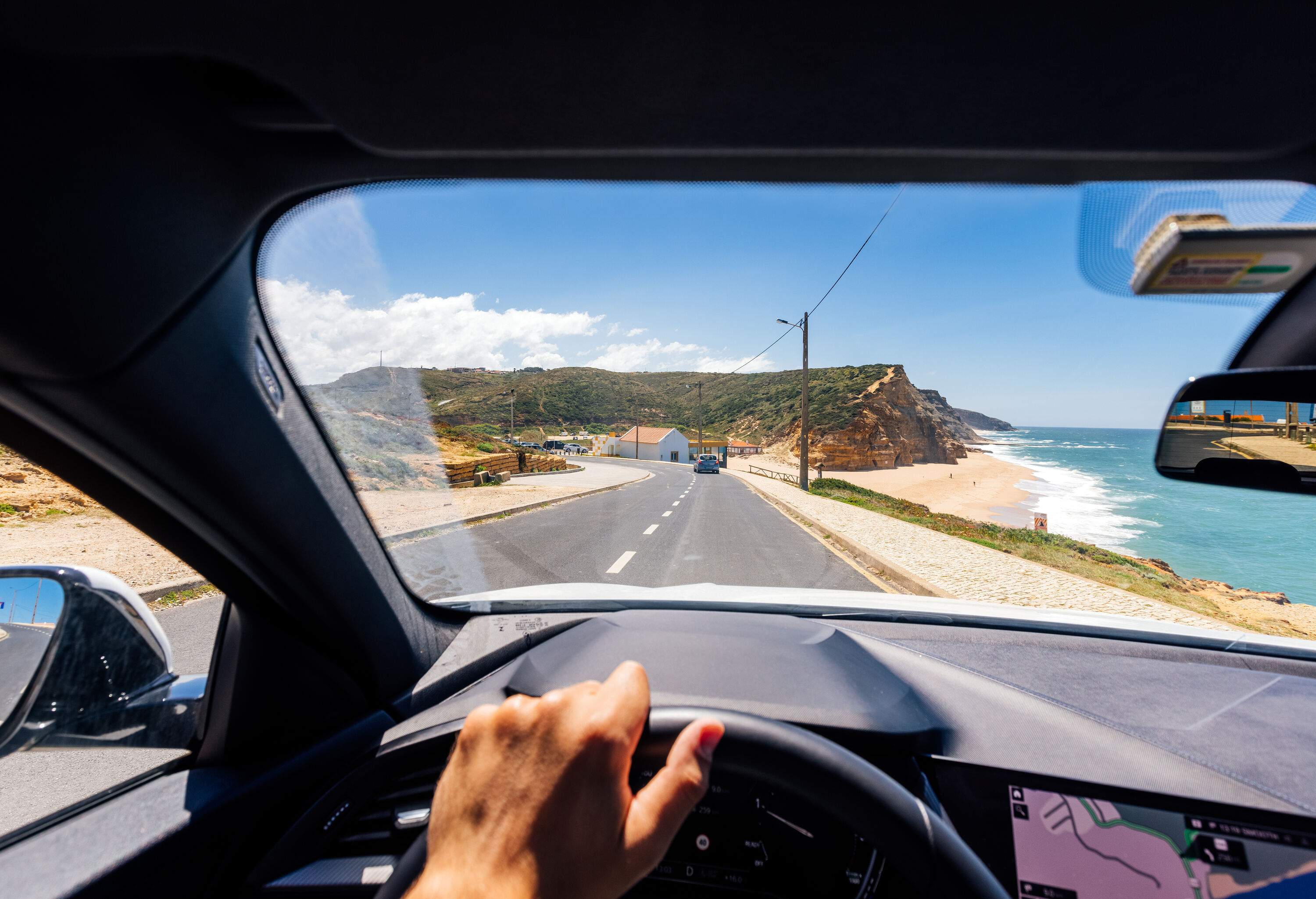 A hand on a car's steering wheel with a front view of a coastal road.