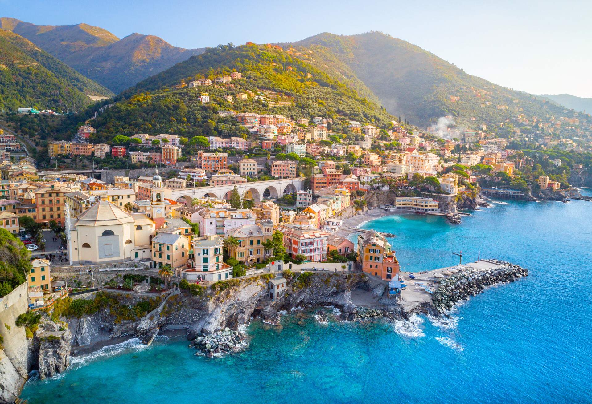 A village with buildings set on steep coastlines at the base of mountains covered in lush foliage.