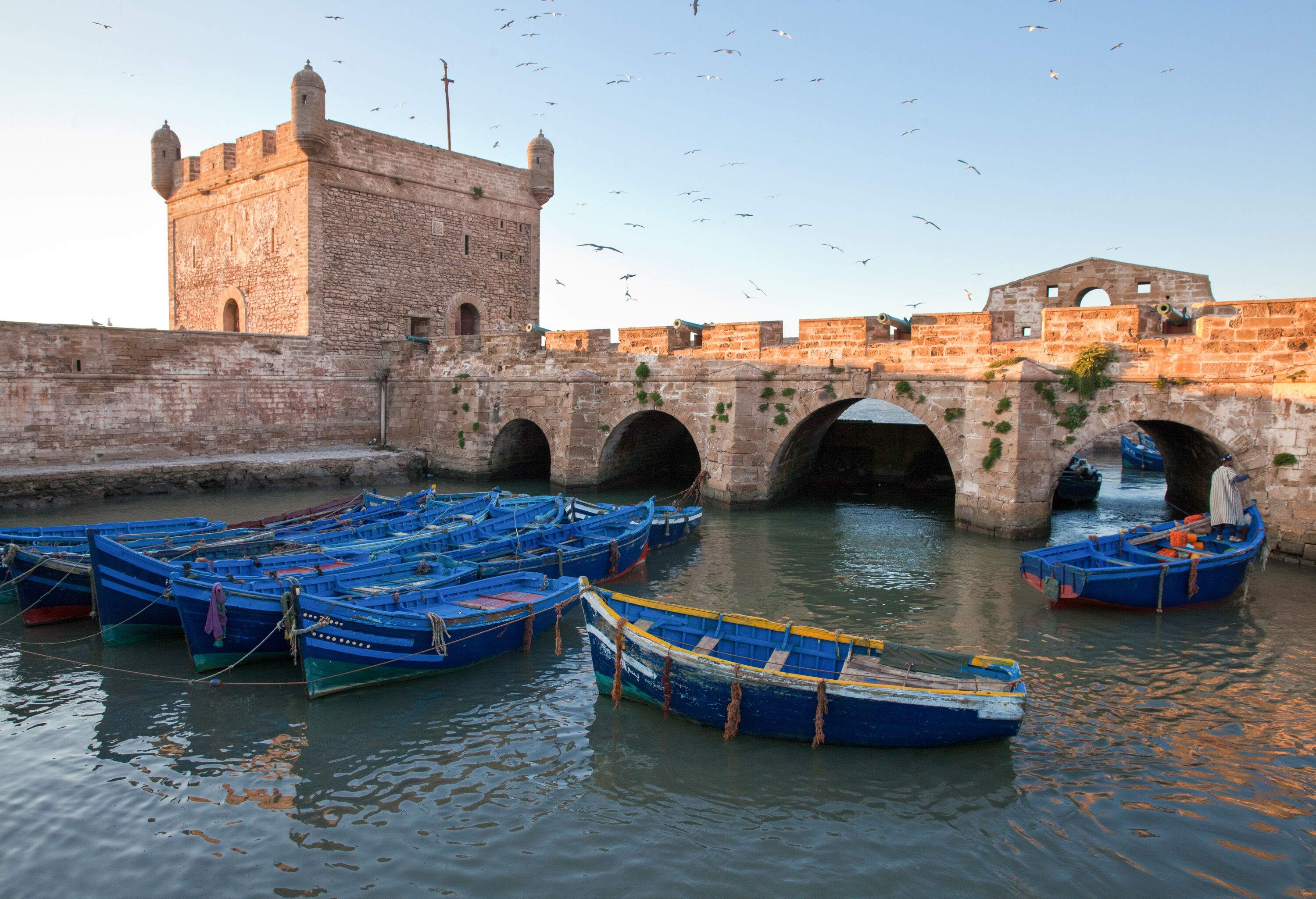 An array of boats of different shapes and sizes are docked against a sturdy stone structure beside a picturesque stone bridge.