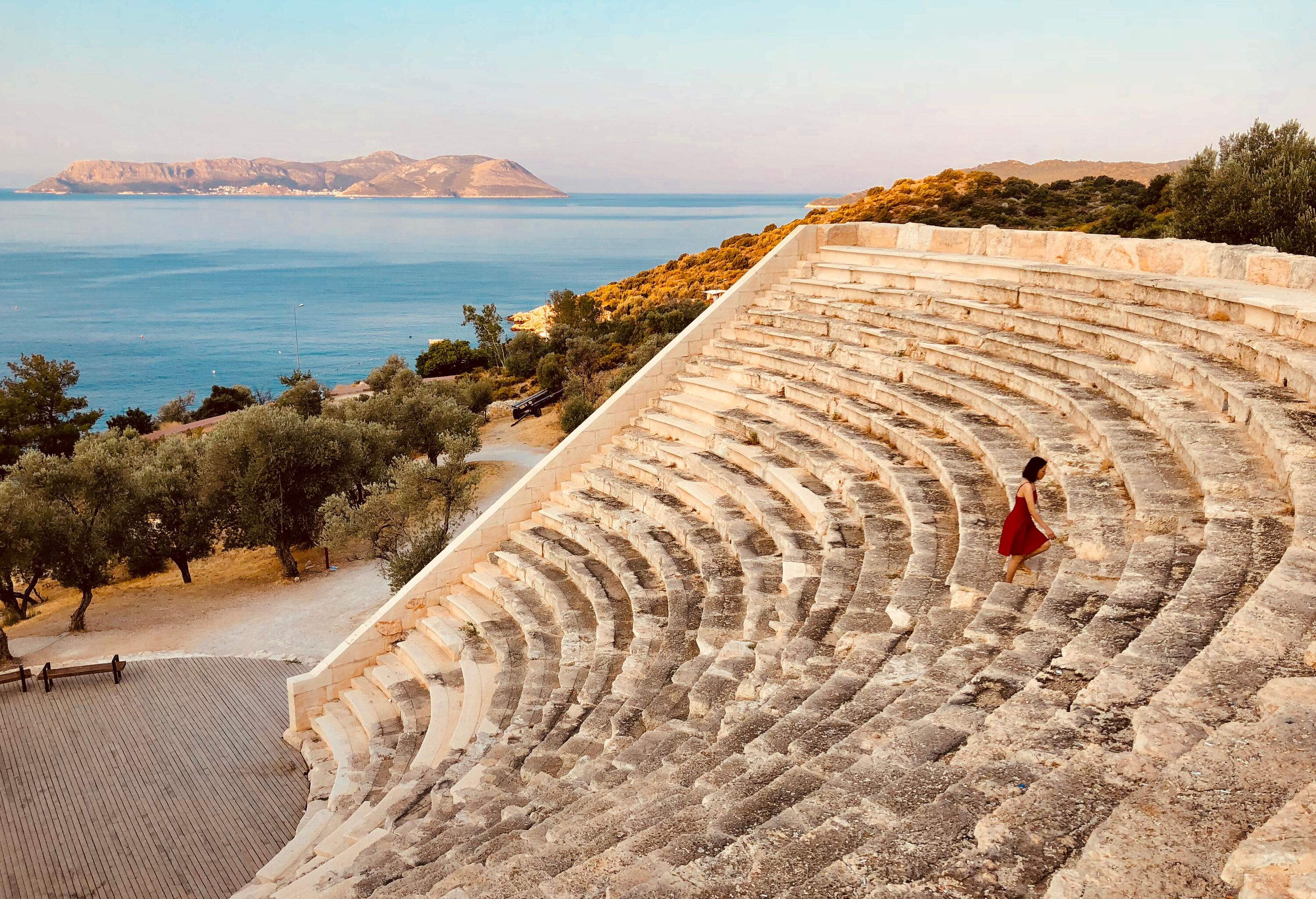A lady tourist wearing a red dress goes up to the ruined amphitheater on a seaside hill.