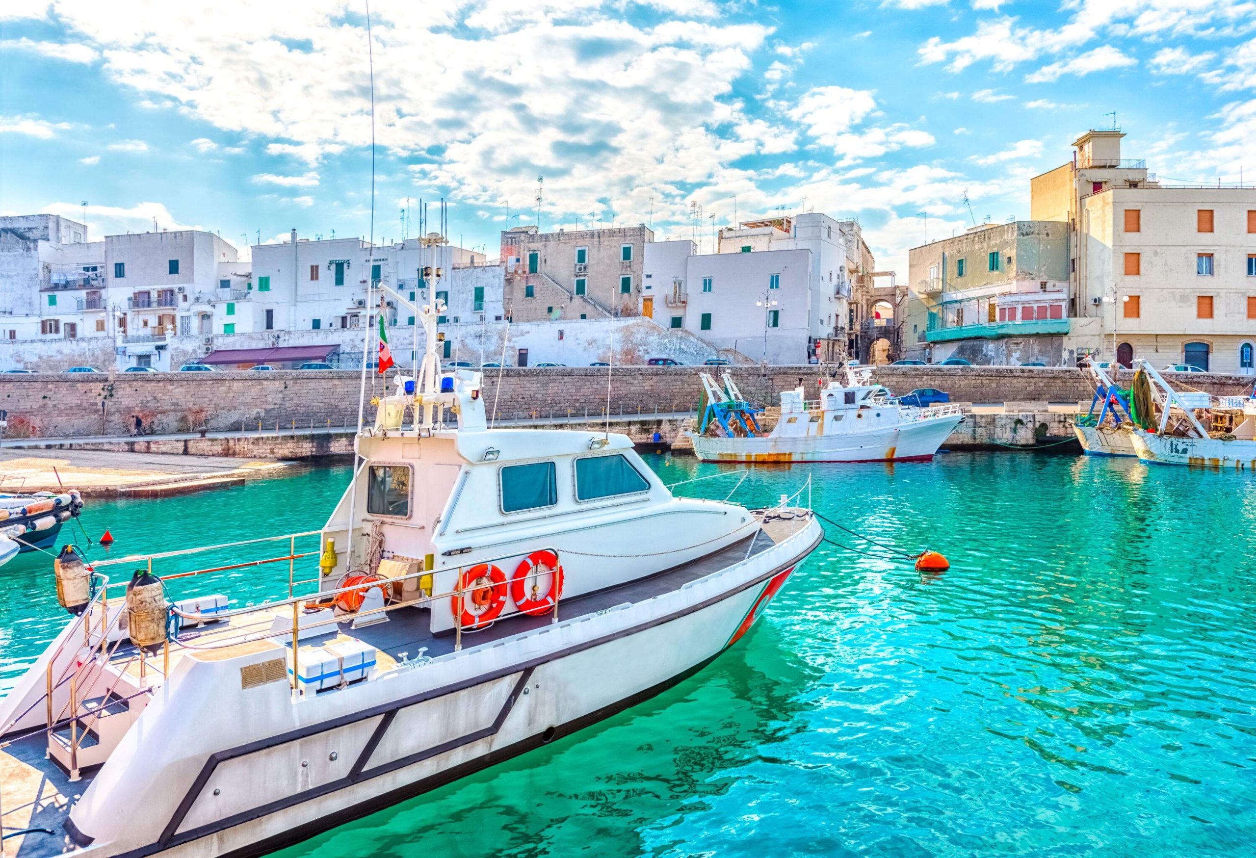 Some yachts and boats moored on a harbour alongside a road lined with parked cars and houses.