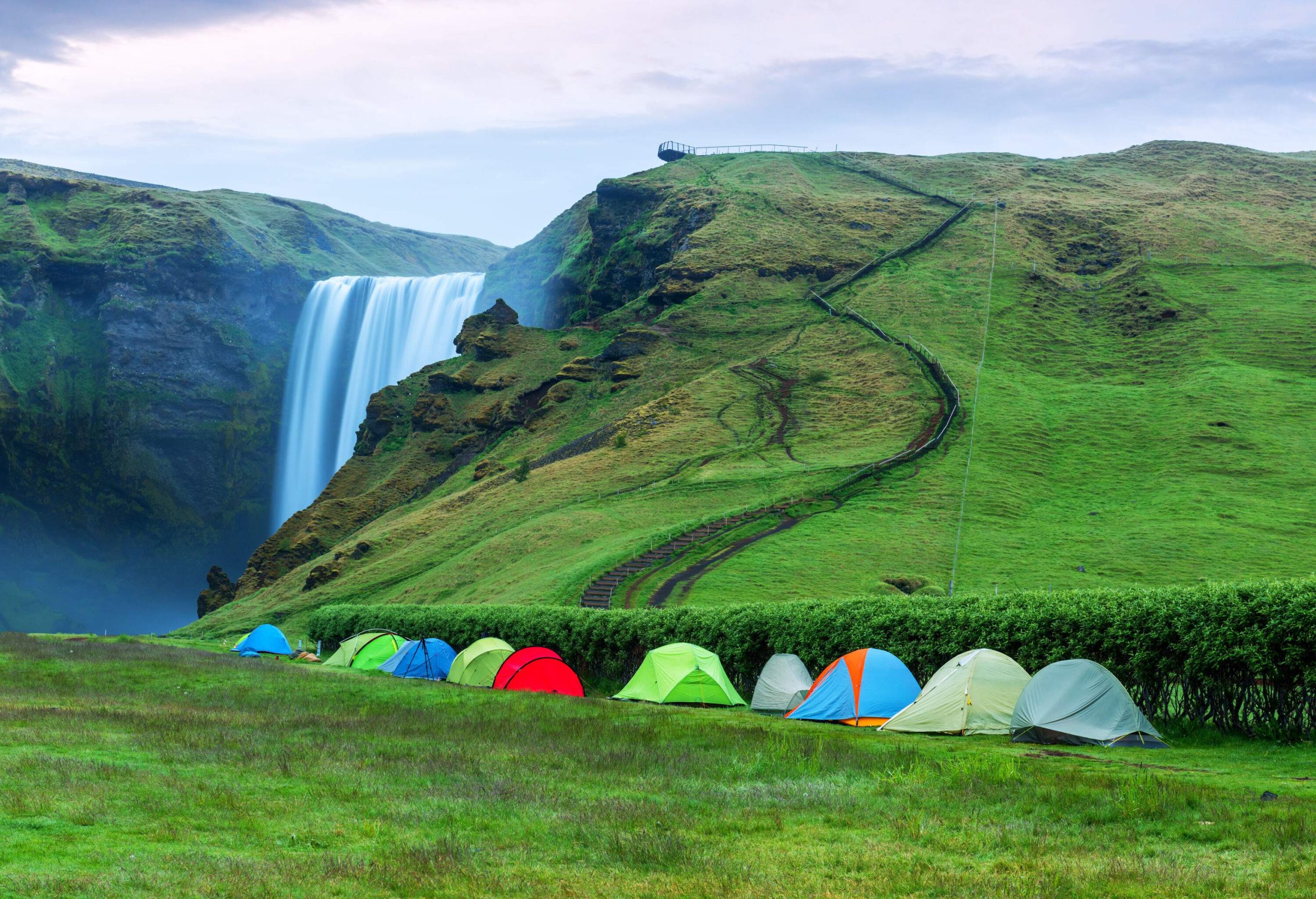 Amidst the lush green surroundings and ascending slope terrain, a picturesque row of colourful camping tents is set up near the iconic Skogafoss waterfall.