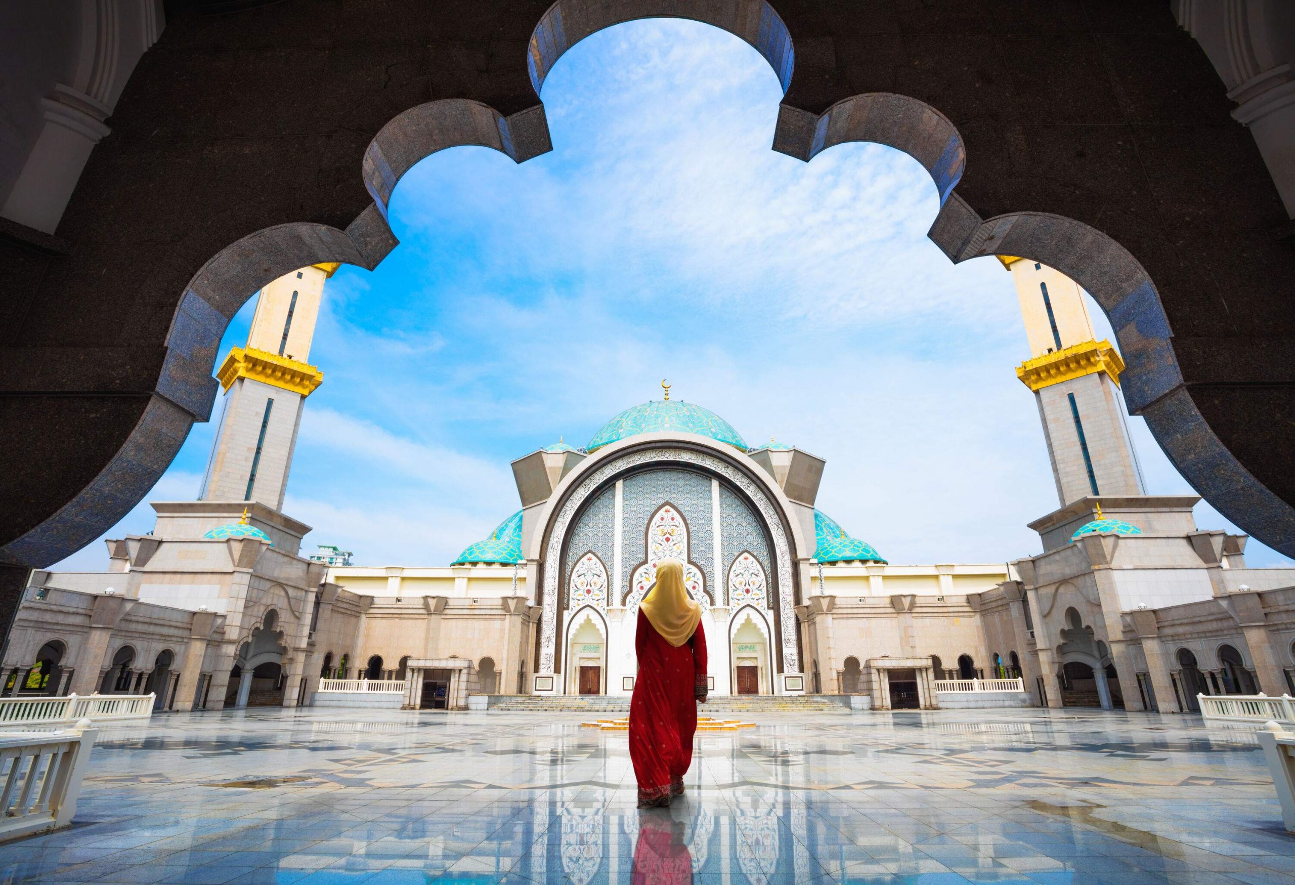 A rear view of a woman in a long red dress with a yellow headscarf walks on the glass-like courtyard of a lovely mosque.