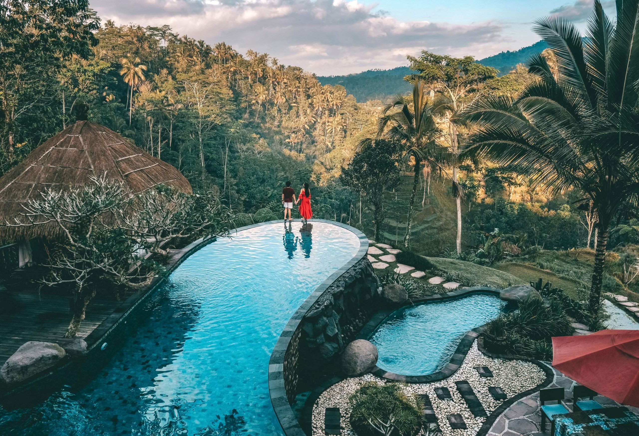 Two people hold hands as they stand at the edge of a luxury resort pool overlooking a lush forest.
