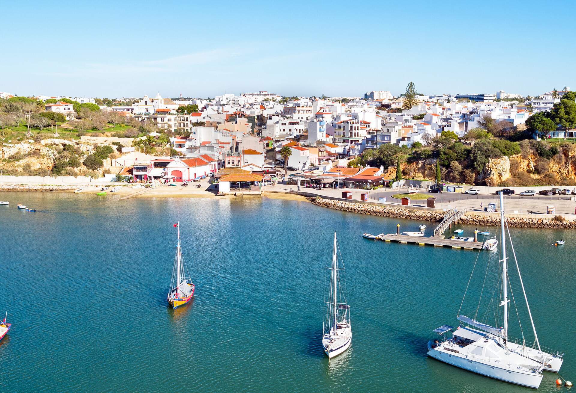 Colourful boats in the sea and a fishing village with white houses surrounded by tall green trees.