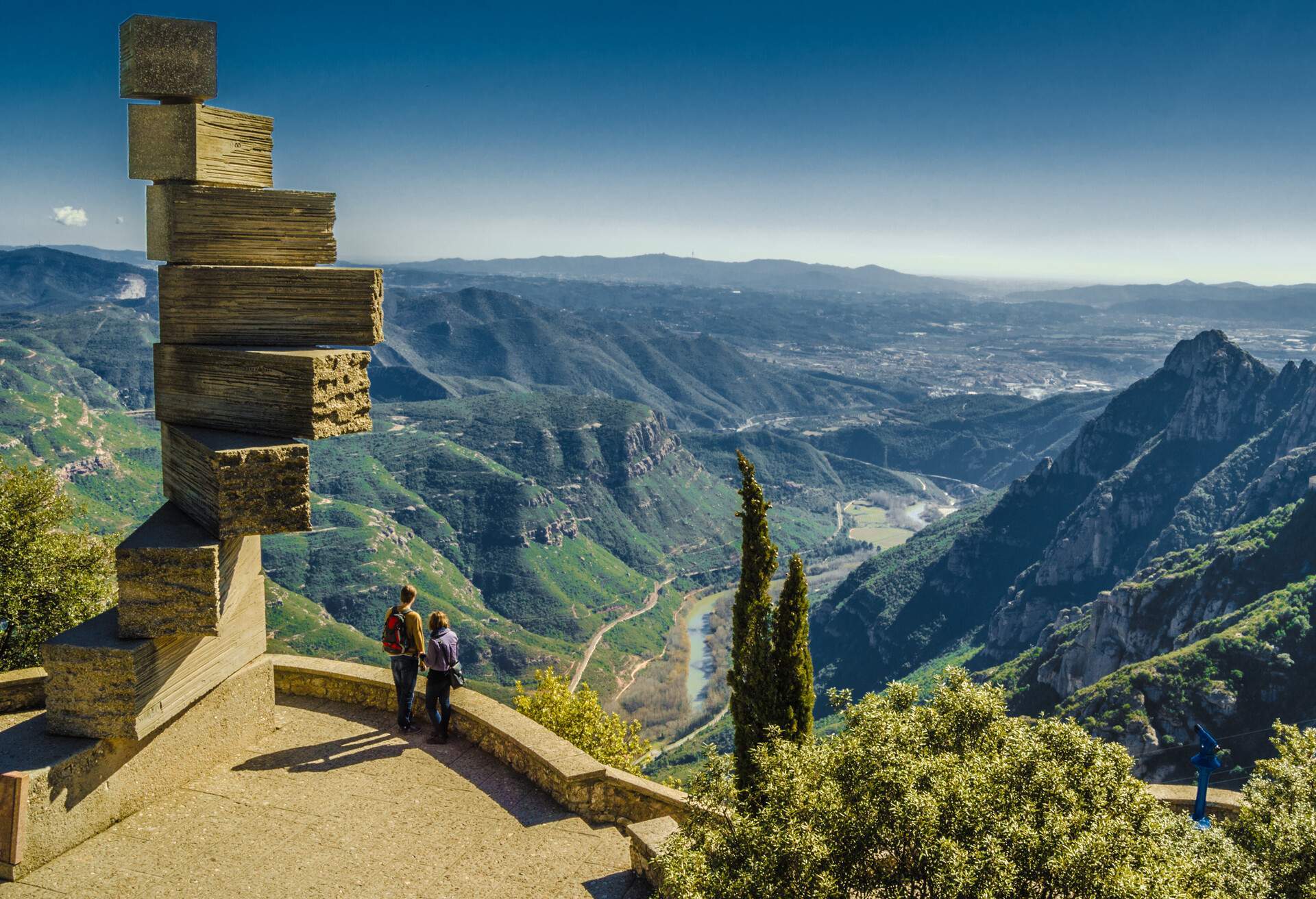 Two people standing on the ledge viewing the mountains with spiral steps of rectangular blocks behind them.