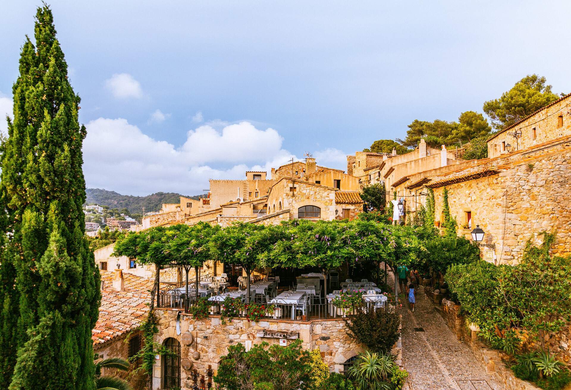 People walking up a steep street past a restaurant and stone houses.