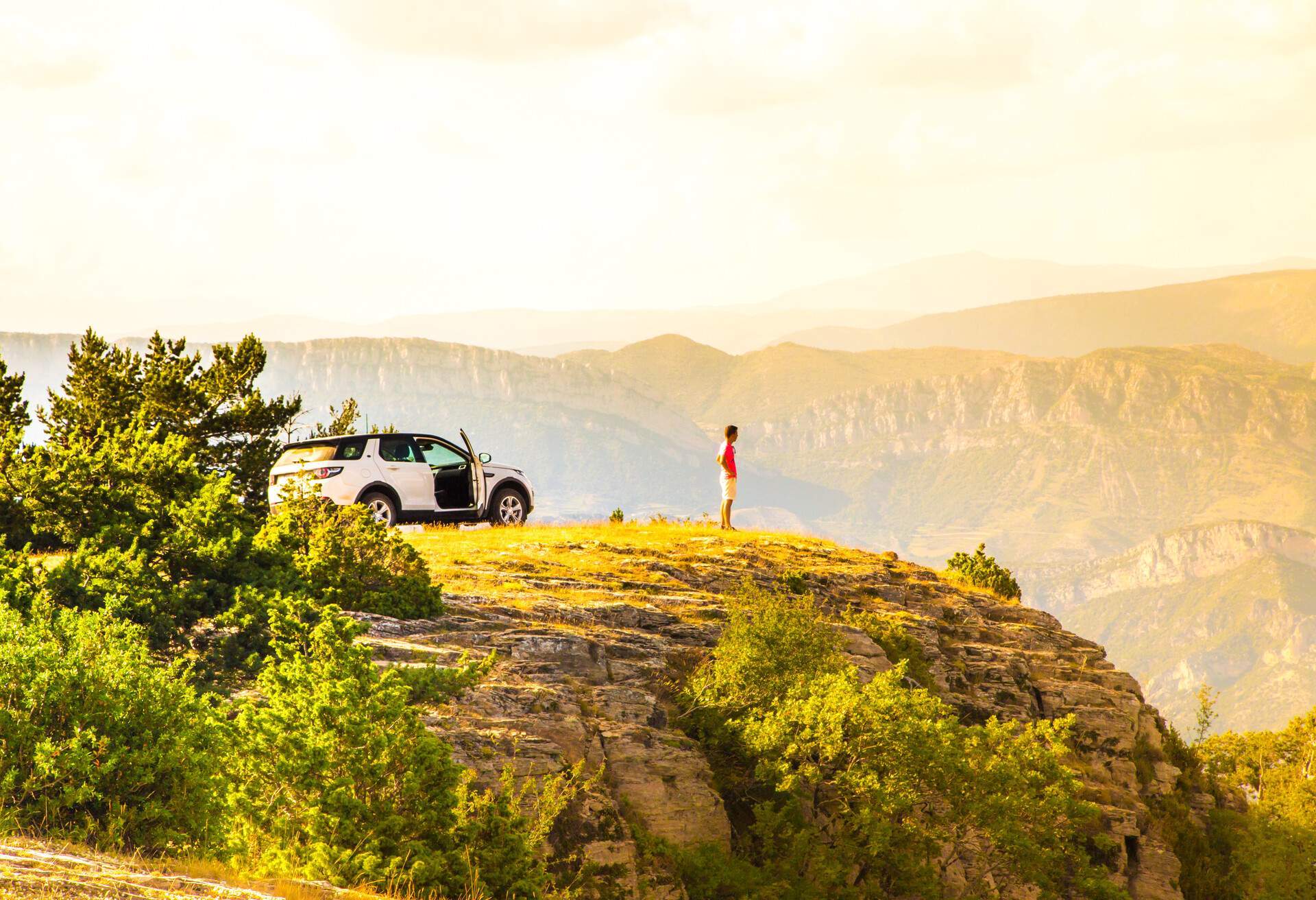 A man standing on a grassy hilltop looking over the valleys and distant mountains.