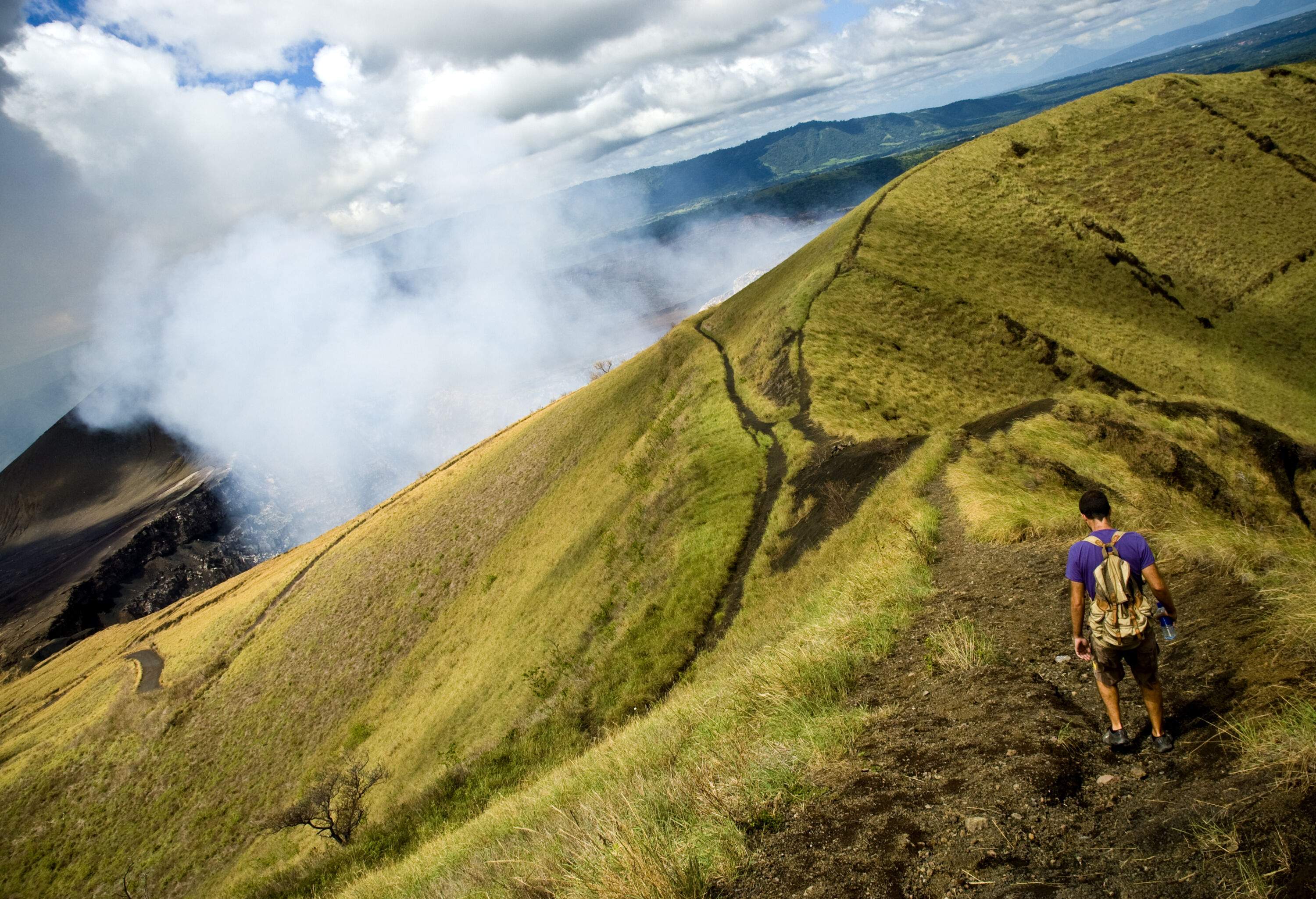 A person walks along a grassy trail near the crater's edge, with wisps of smoke rising from the depths.