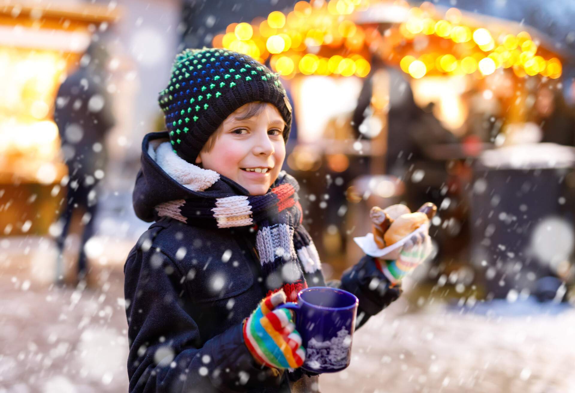 Petit garçon souriant mangeant une saucisse allemande et buvant du punch chaud pour enfants sur le marché de Noël. Enfant joyeux sur le marché familial traditionnel en Allemagne. Garçon rieur en vêtements d'hiver colorés.
