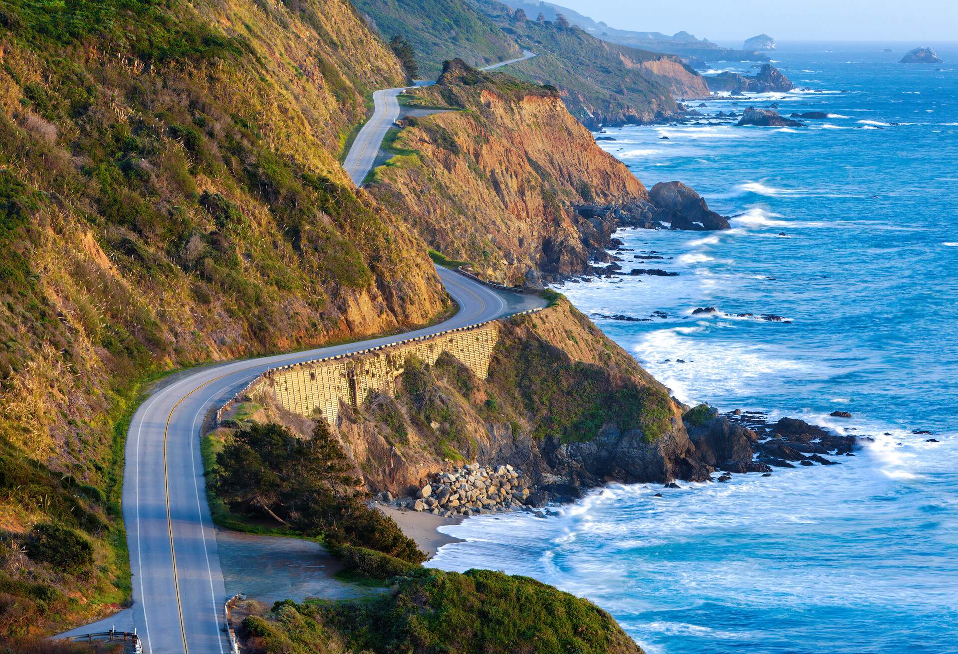 A winding road with views of a rocky beach carved into the faces of high coastal cliffs.