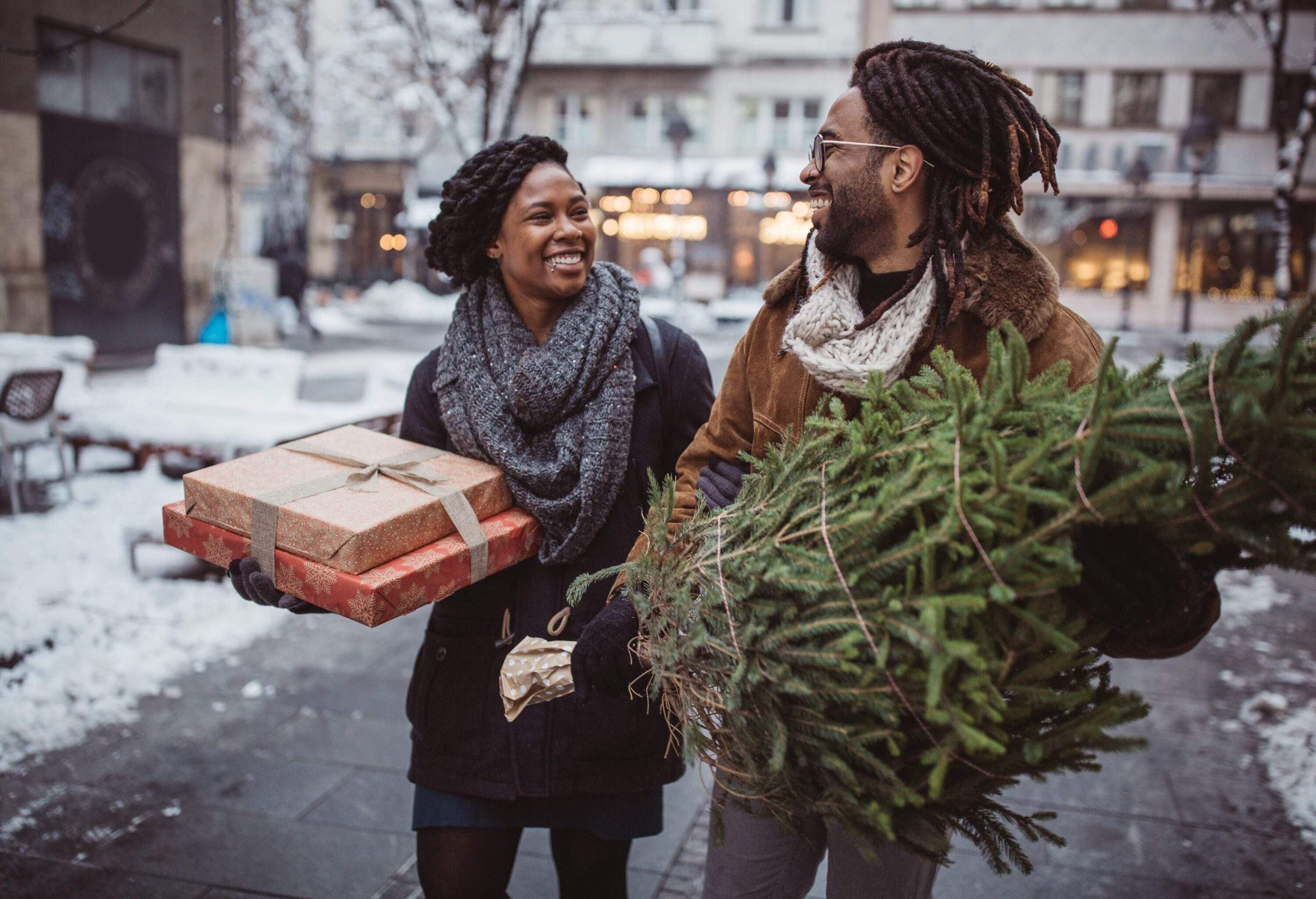 Un couple en vêtements d’hiver se sourit en portant des cadeaux emballés et un arbre de Noël.