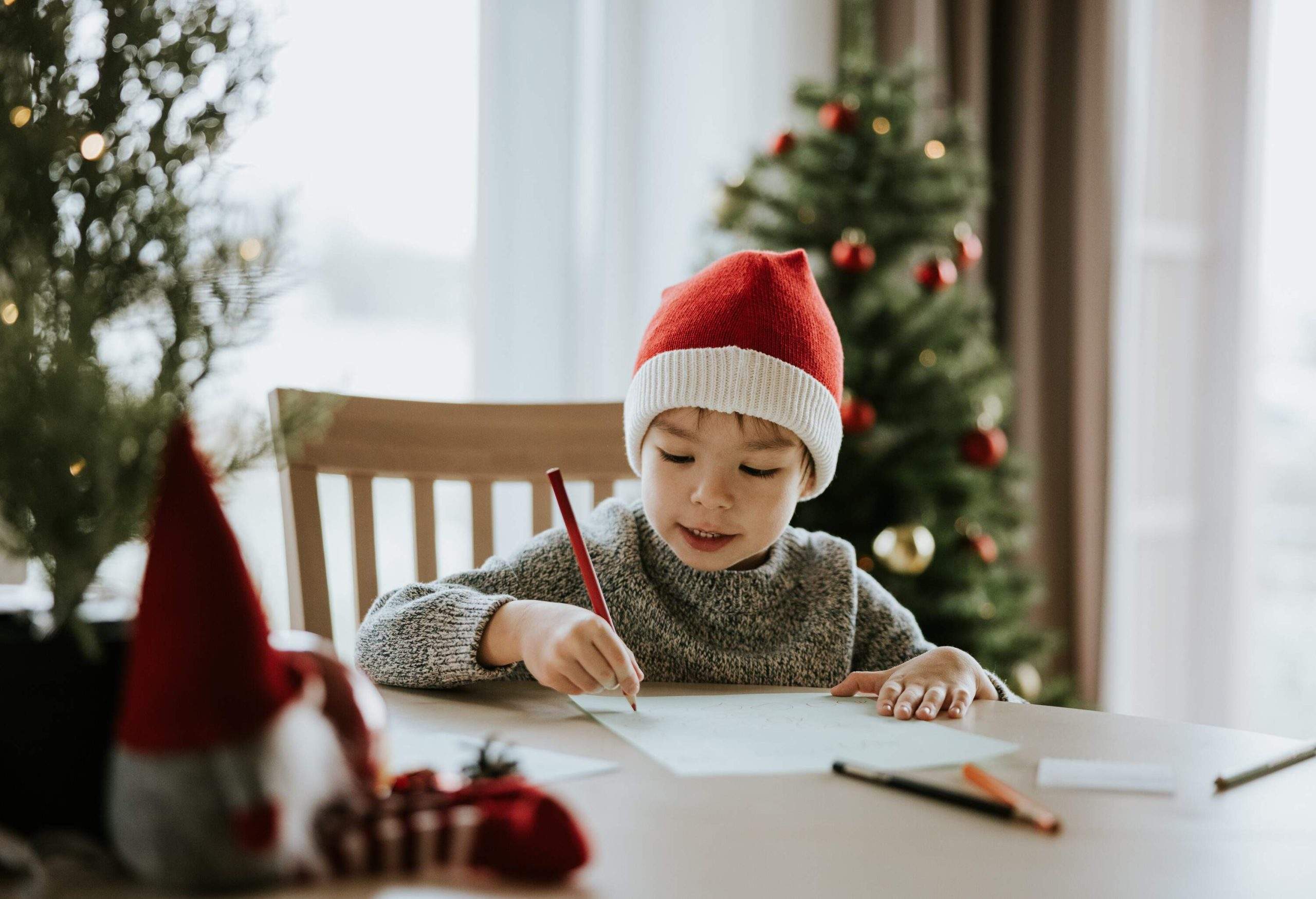 Un petit garçon assis à une table en train d'écrire une lettre, portant un bonnet de Père Noël