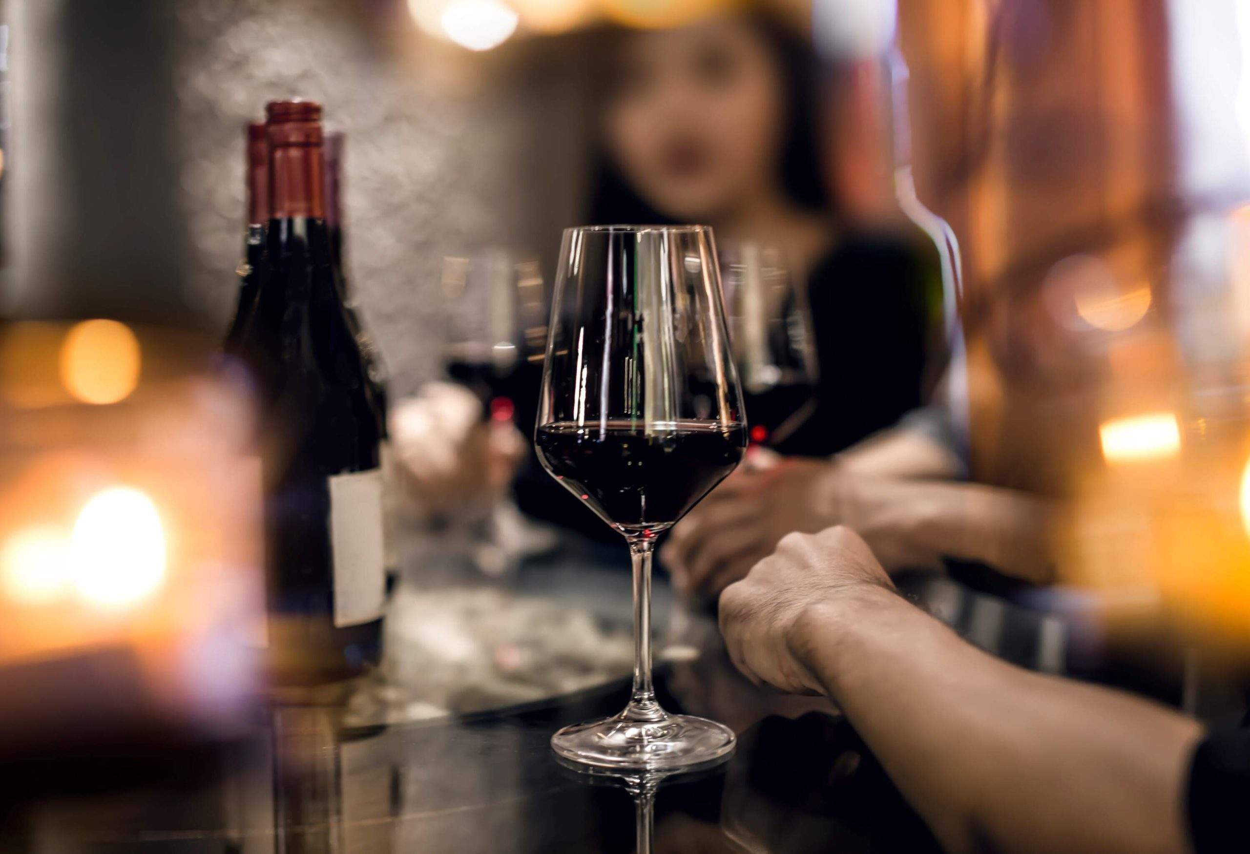 Un verre de vin rouge est posé sur une table, entouré d’un groupe d’amis en arrière-plan, créant un effet flou.