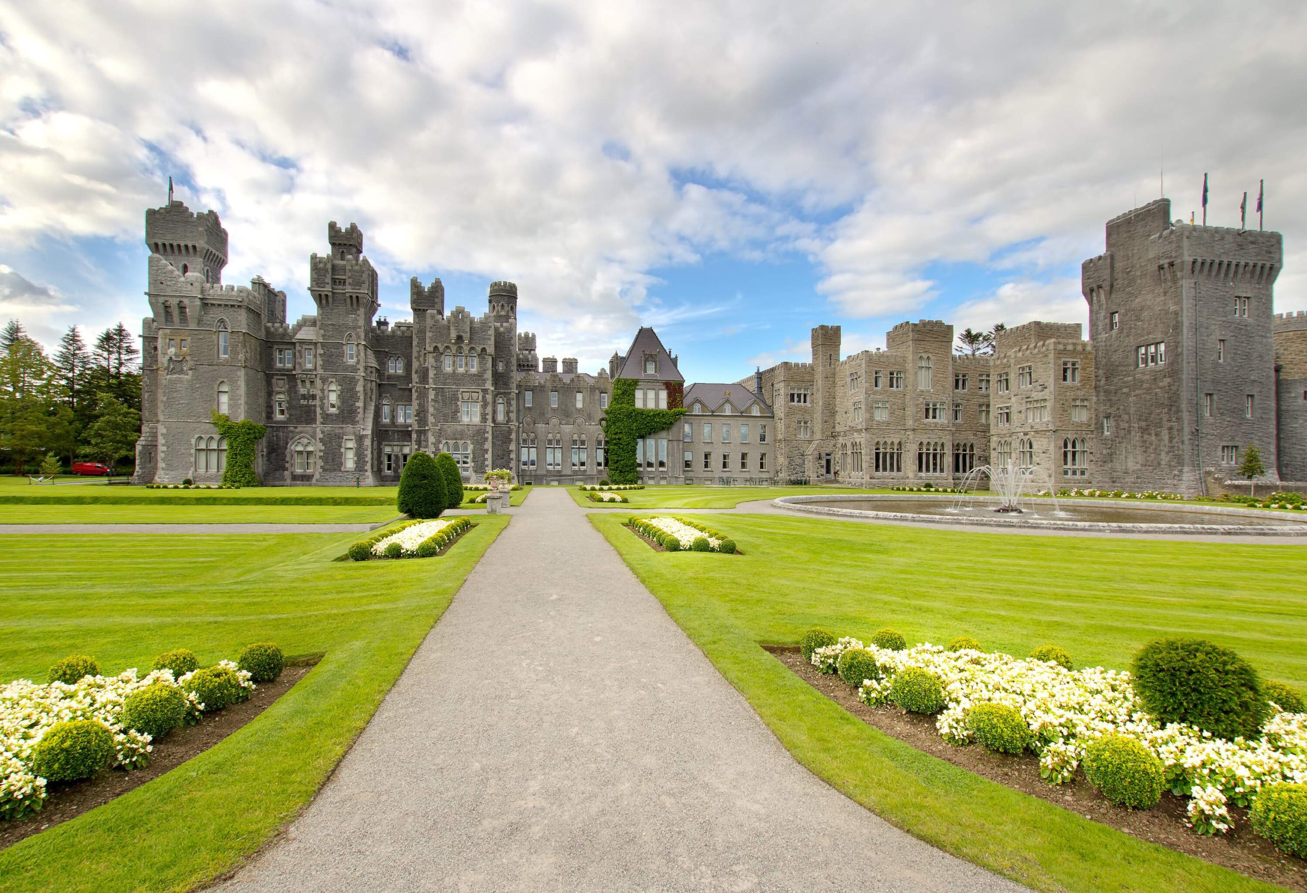 The landscape of a medieval and Victorian architecture castle with a lush green meadow in front and a fountain.