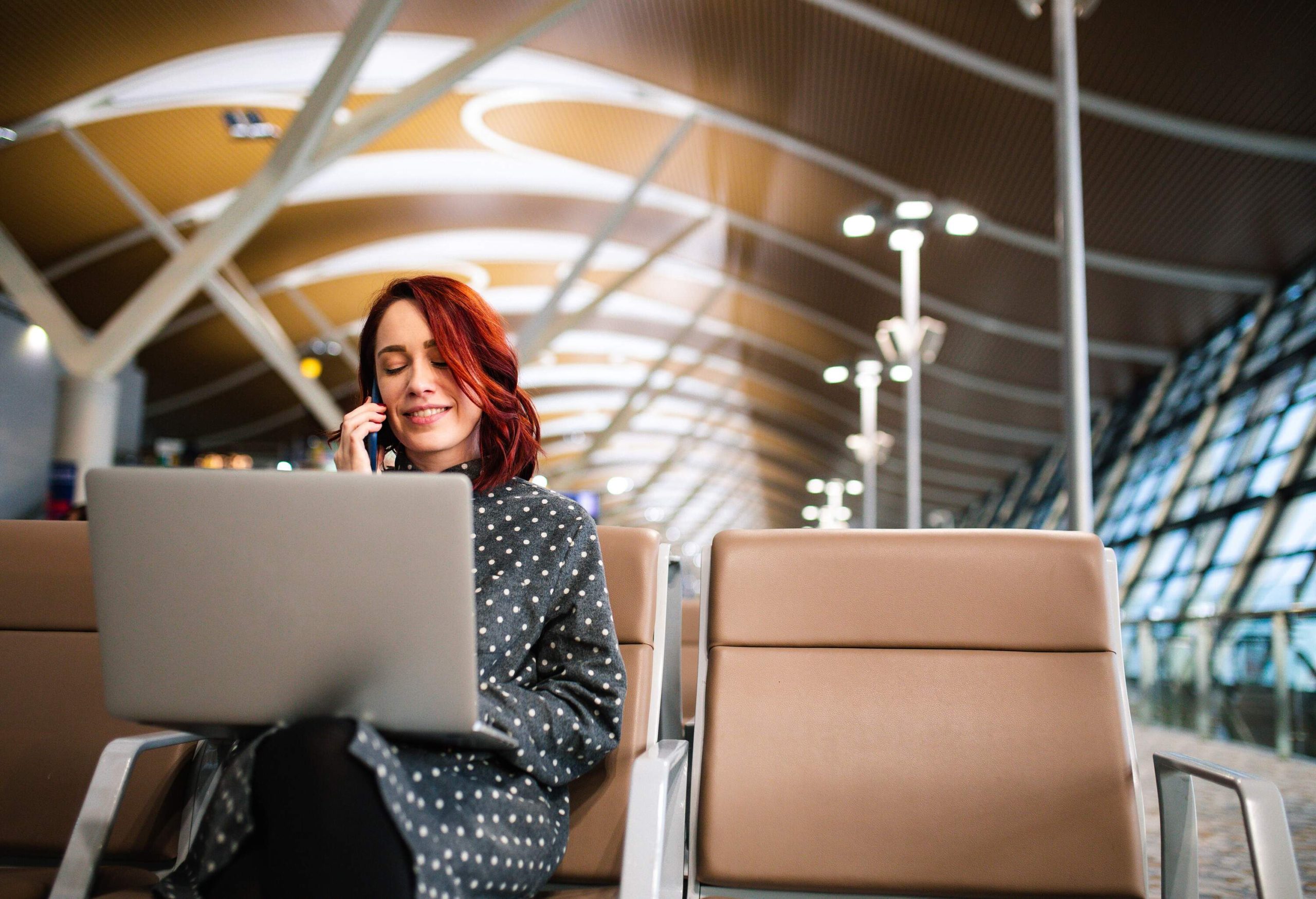 A woman sitting in an airport terminal talking on her phone while holding her laptop in her lap.