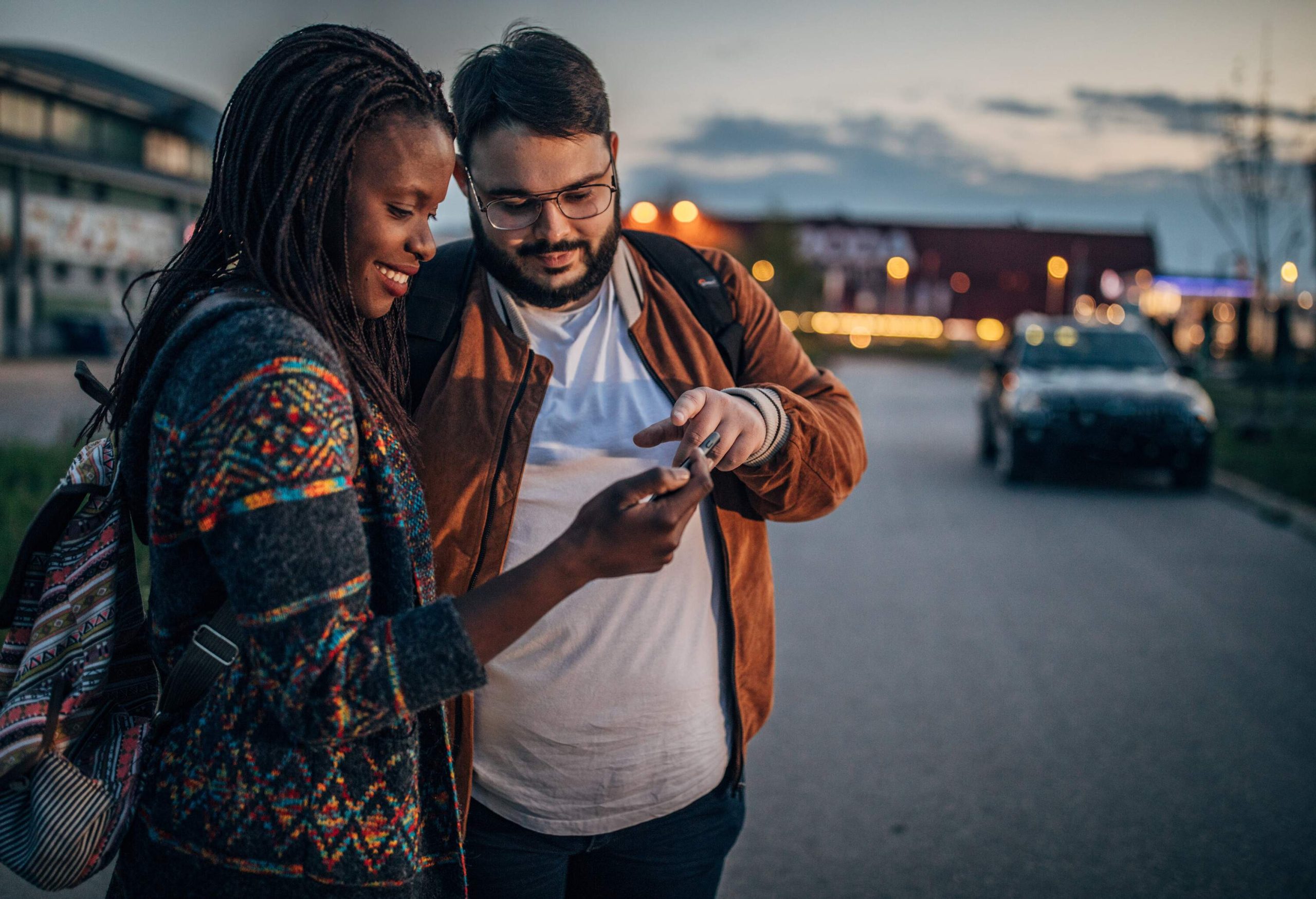 A man and a woman checking on a smartphone while standing on the side of a road.