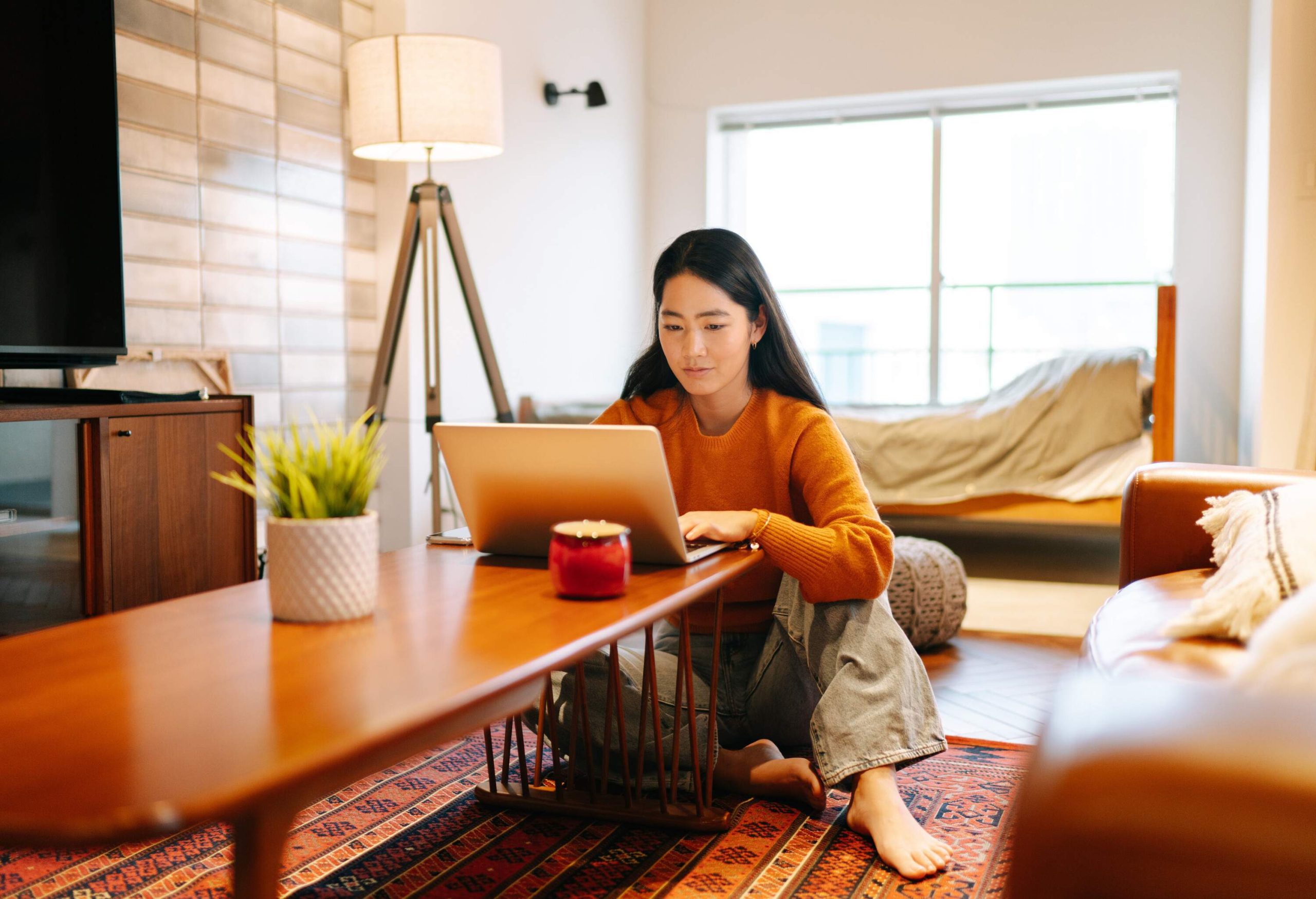 A black-haired woman in an orange sweater sits on the living room as she uses a laptop on the coffee table.