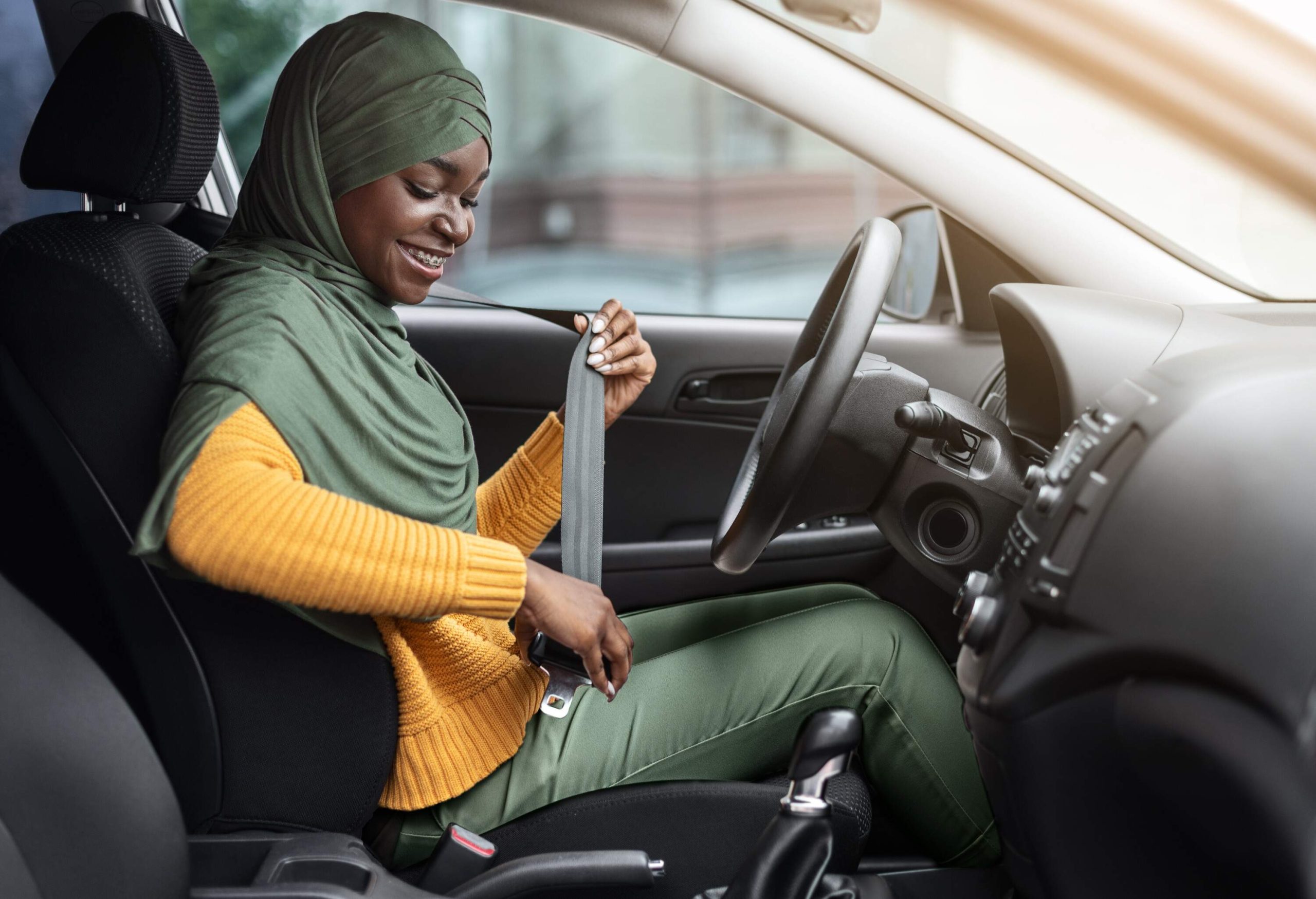 A woman wearing a hijab and a yellow sweater smiling as she buckles her seatbelt in the driver's seat.