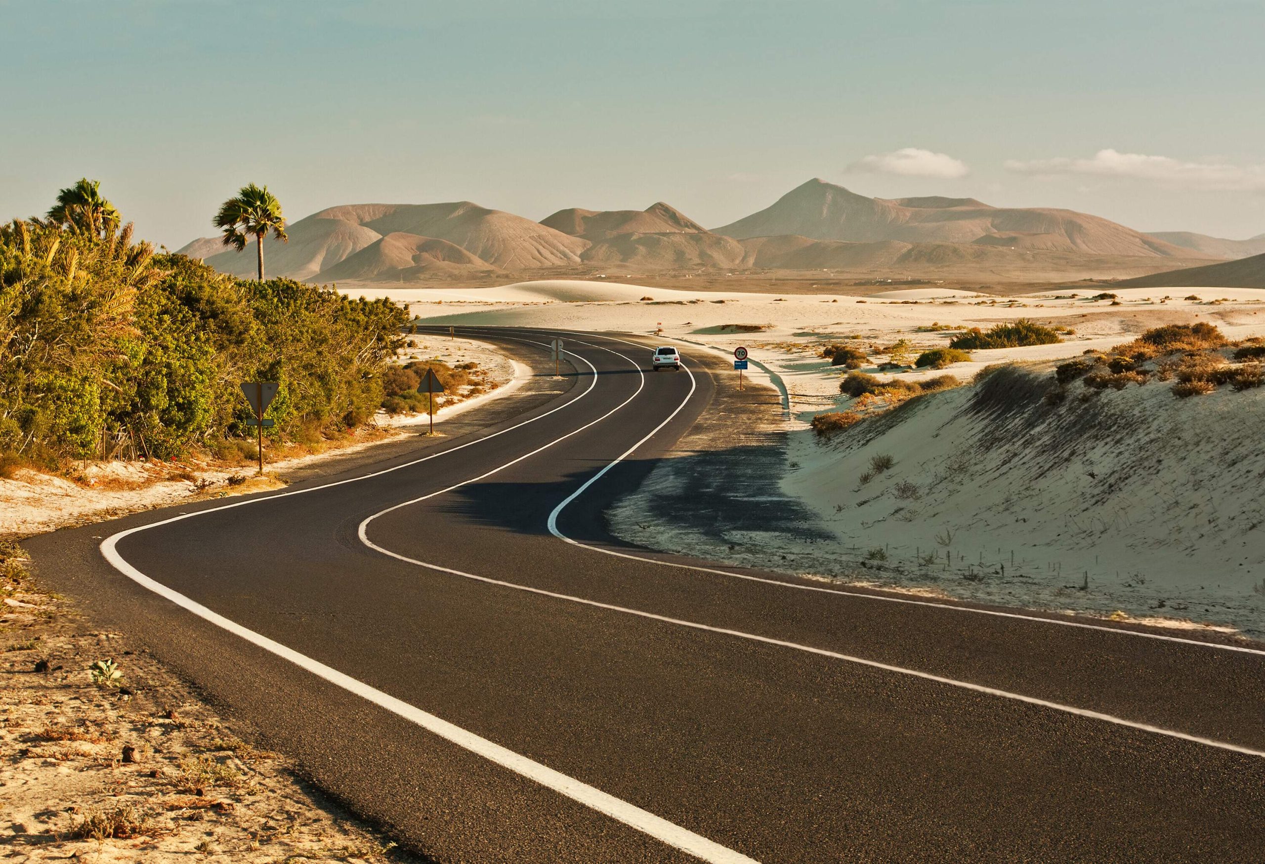 A car travels down a winding road surrounded by grassy sand dunes with distant mountain views.