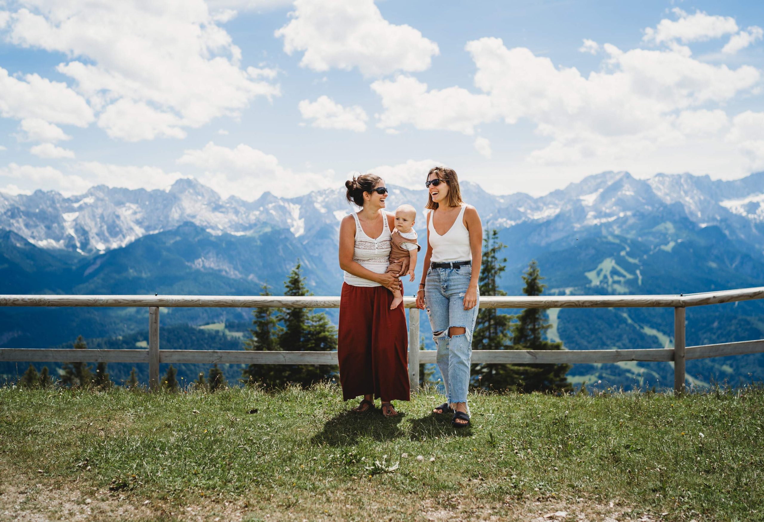 Two women with a baby standing on a fenced-off highland with craggy mountains behind them.