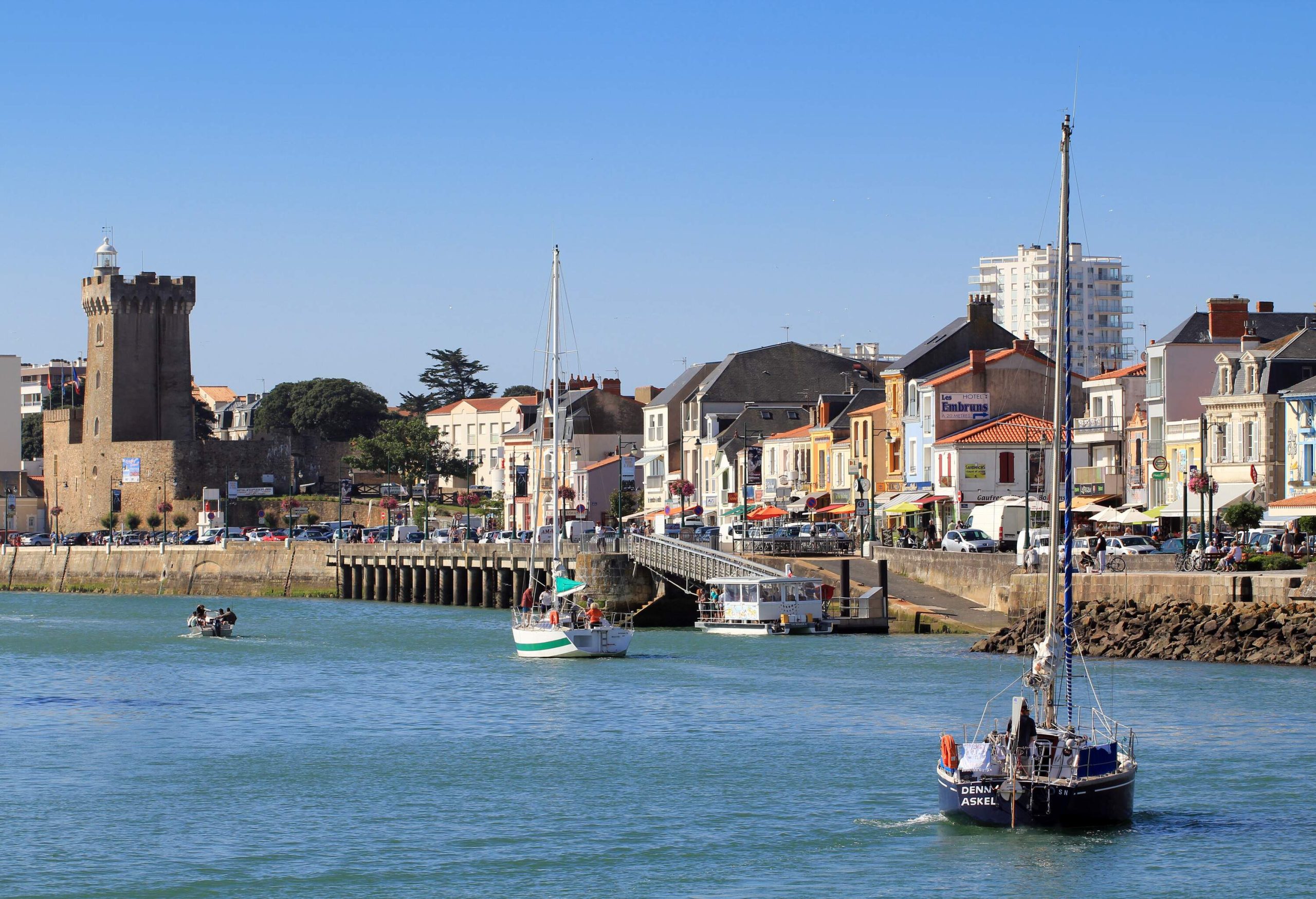 Boats pass in front of a seaside town with a tower made of brick that looks out over the water.