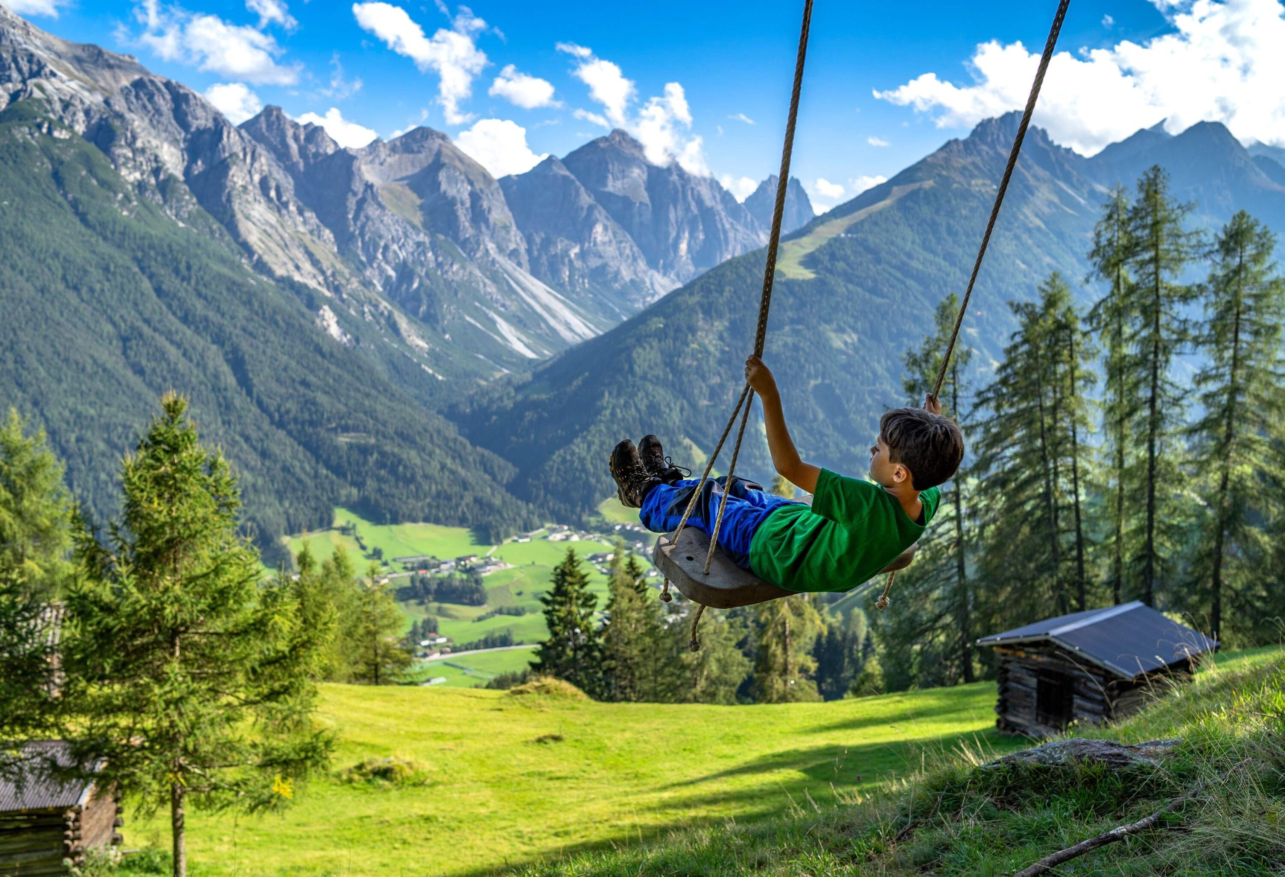 A young boy in a green shirt playing on a swing with views of lush meadows and craggy mountains.