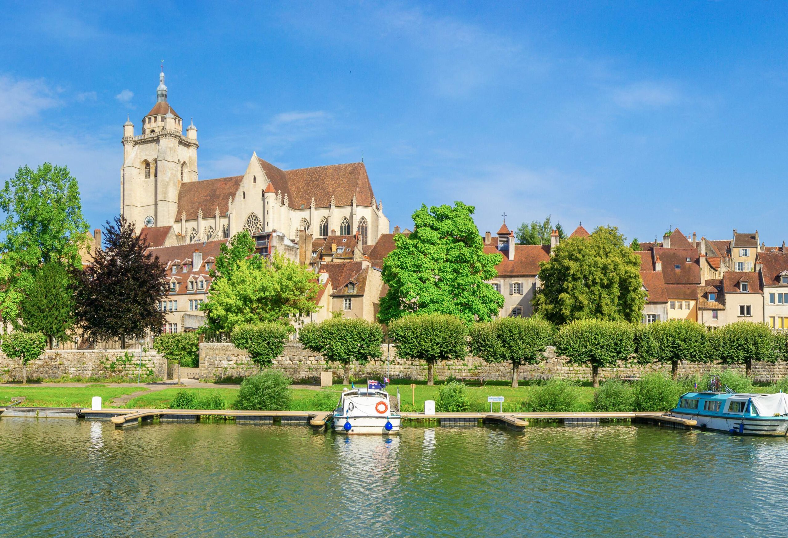 A small village dominated by a church alongside a canal with docked boats and trees on its bank.