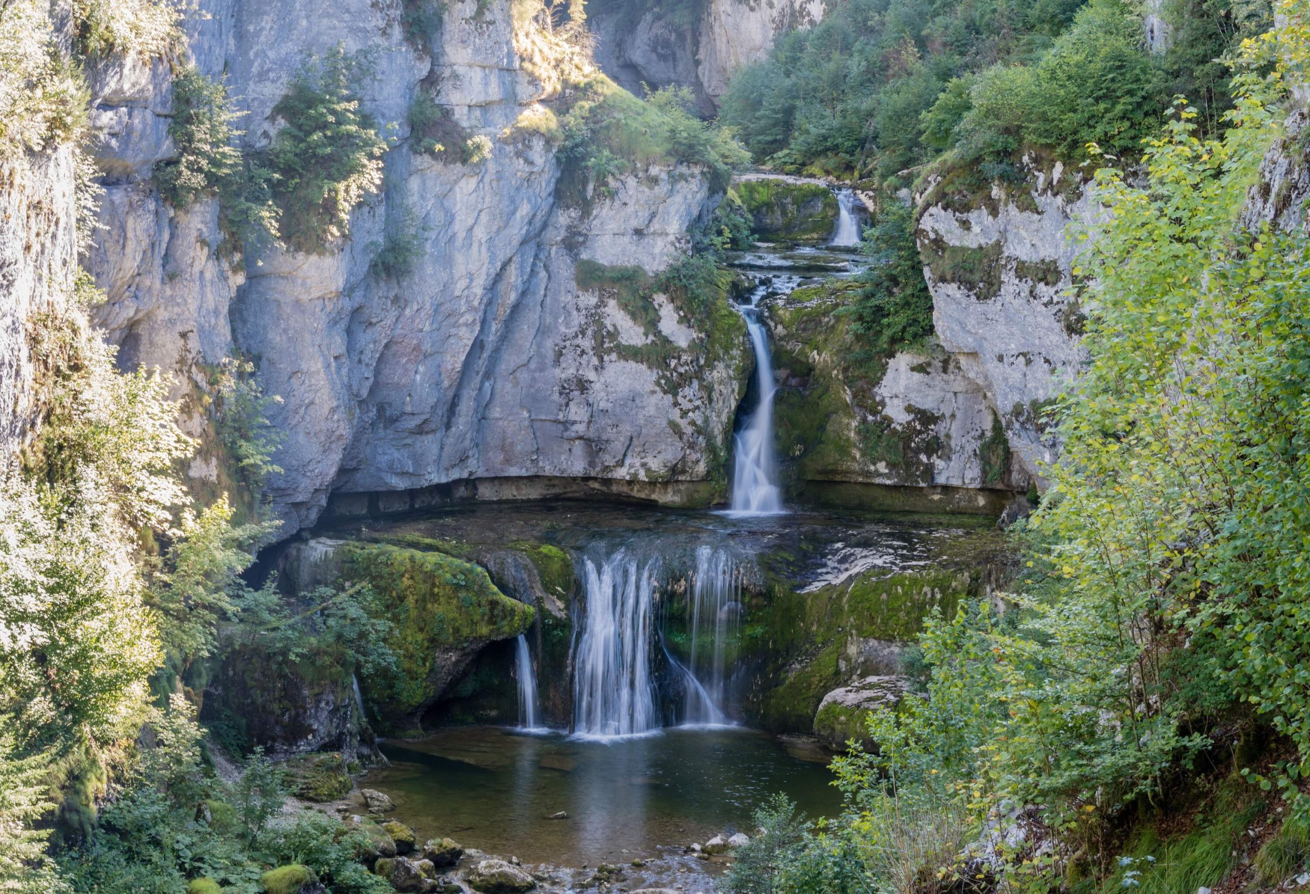 As the river crashes through a tight cleft in a big rock formation, it creates two waterfalls that are surrounded by green plants.