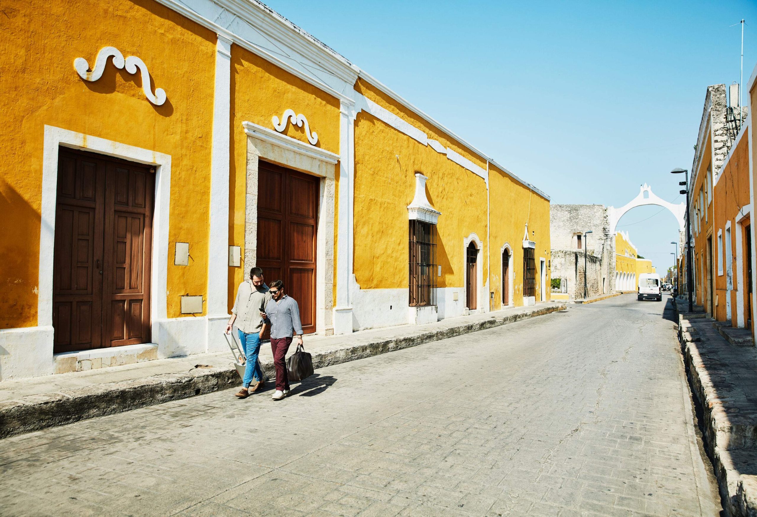 Two men carrying luggage are walking down a cobblestone street lined with yellow-painted houses.