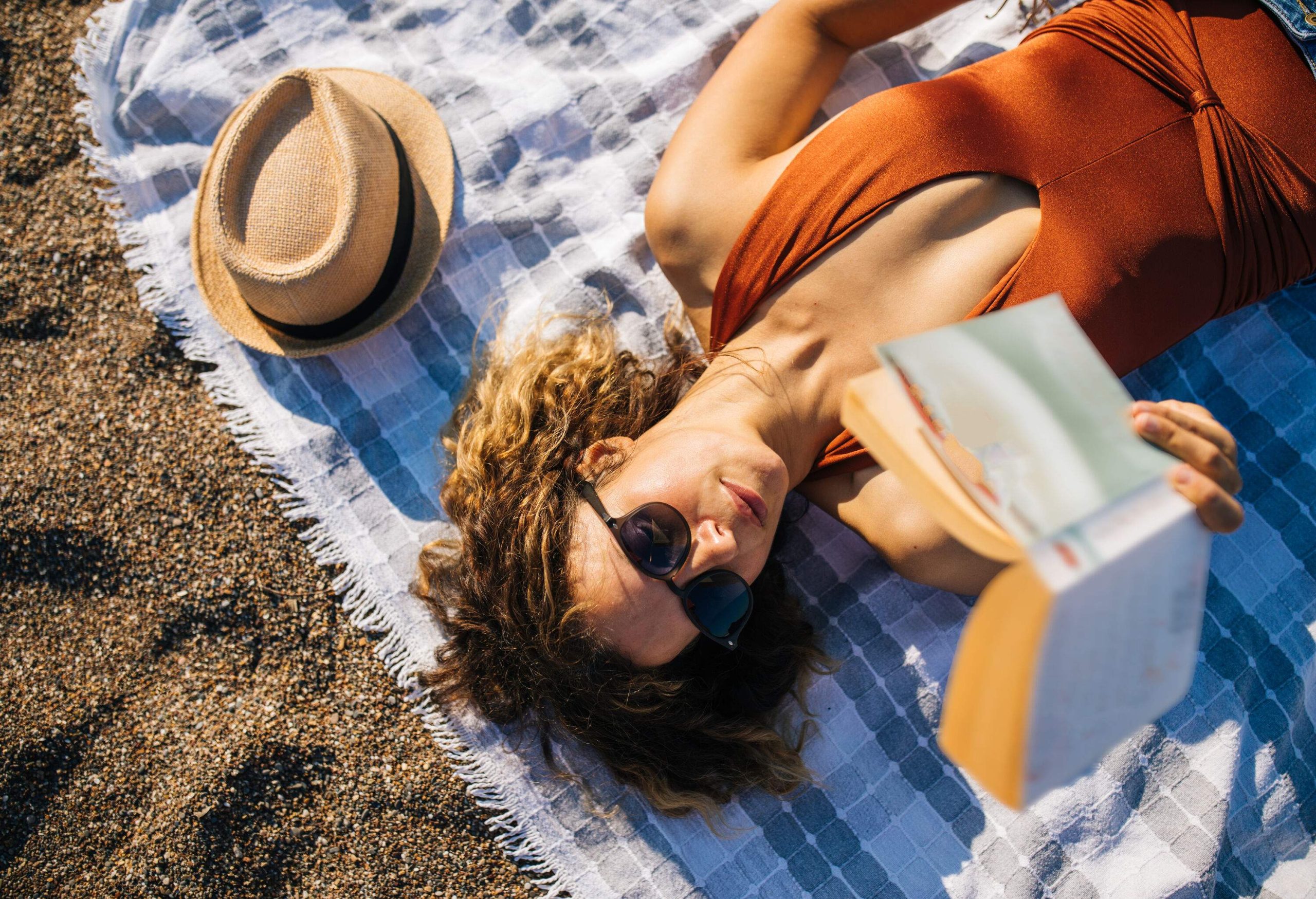 A brunette woman in sunglasses and brown swimwear lies on a picnic blanket beside a straw hat while reading a book.